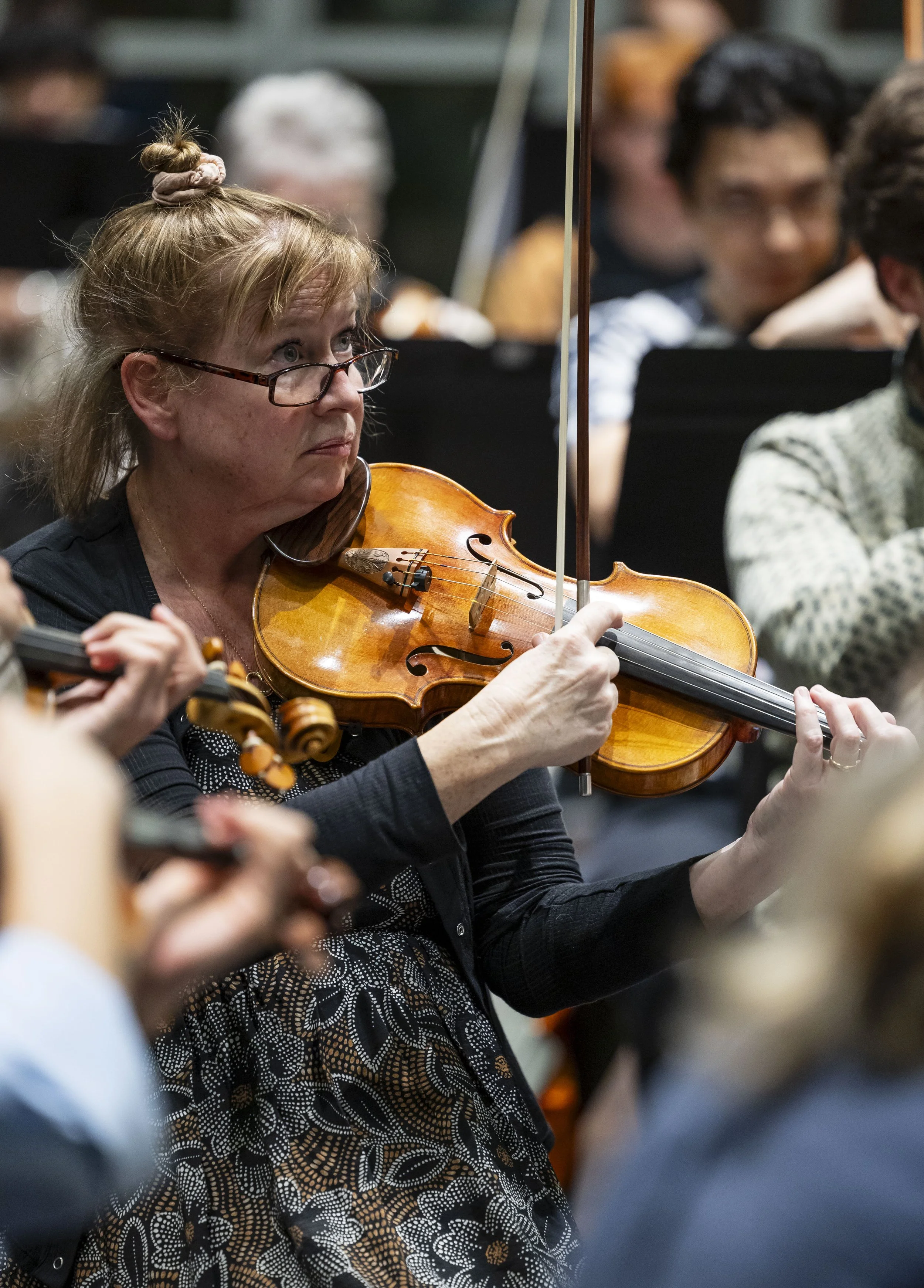 A woman with glasses playing a violin in an orchestra setting, surrounded by other musicians