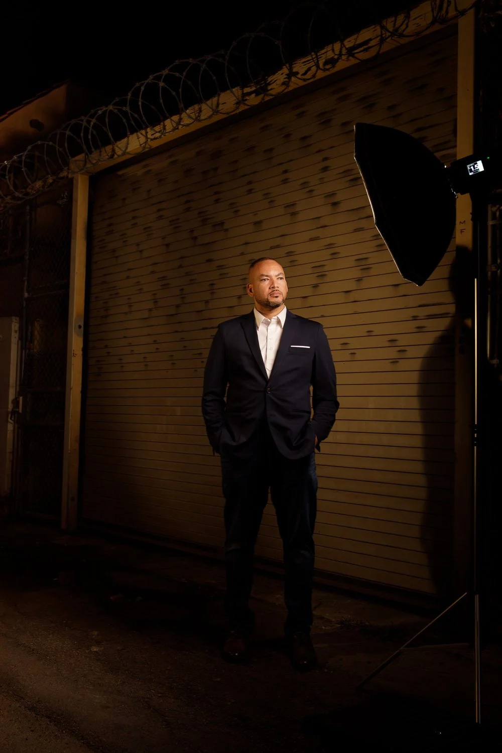 A man wearing a black suit and white shirt stands in front of a wooden garage door at night, with an amber glow illuminated by a large studio light.