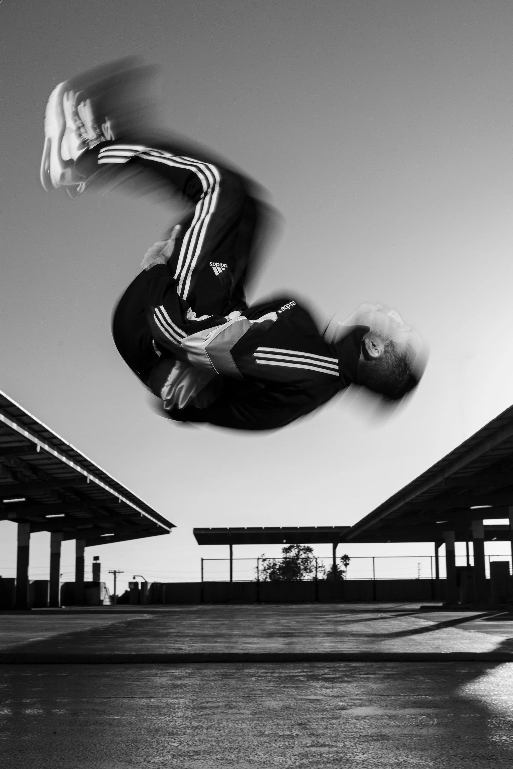 A break dancer in motion captured in a mid-air flip at a parking lot, wearing athletic clothing including a jacket and Adidas sneakers, with a clear sky in the background.