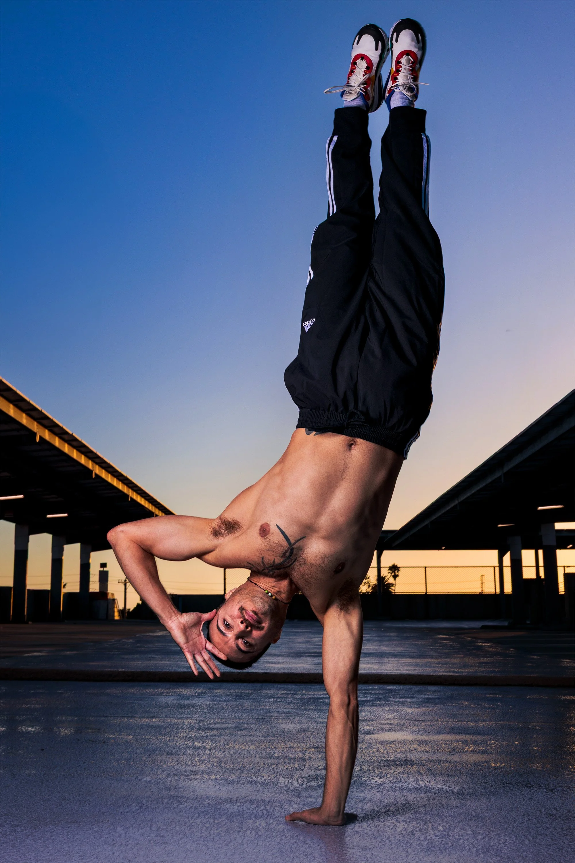 A shirtless man performing a one-handed handstand outdoors at sunset, wearing black athletic pants and colorful sneakers.