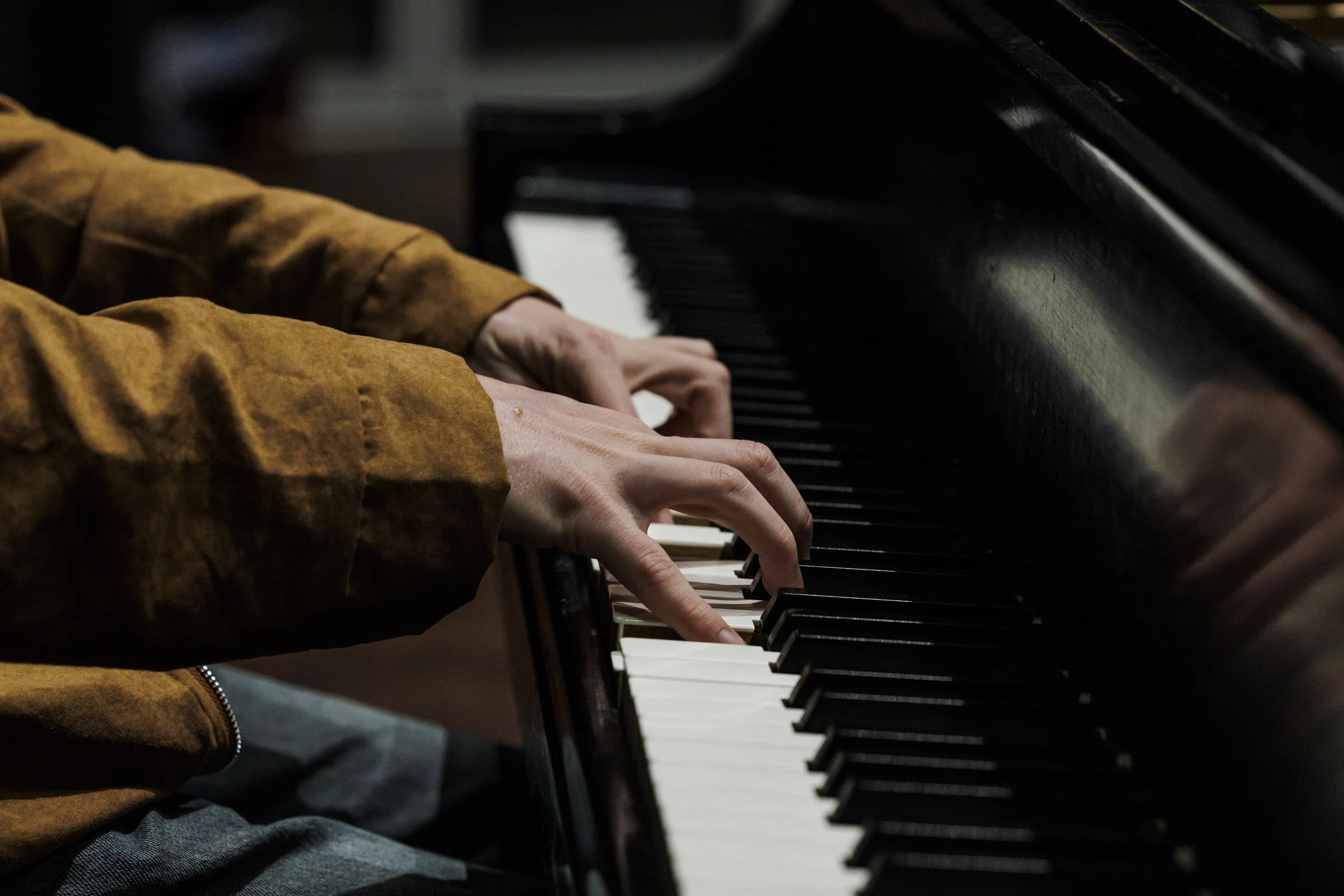 Close-up of a person's hands playing the piano keyboard, wearing a brown jacket.