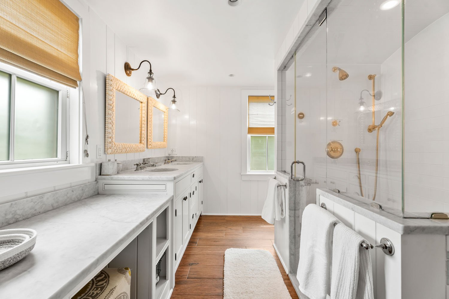 Bright bathroom with white walls, wood flooring, double vanity with marble countertop, window with window shades, shower with glass door, white towels, and wall-mounted light fixtures.