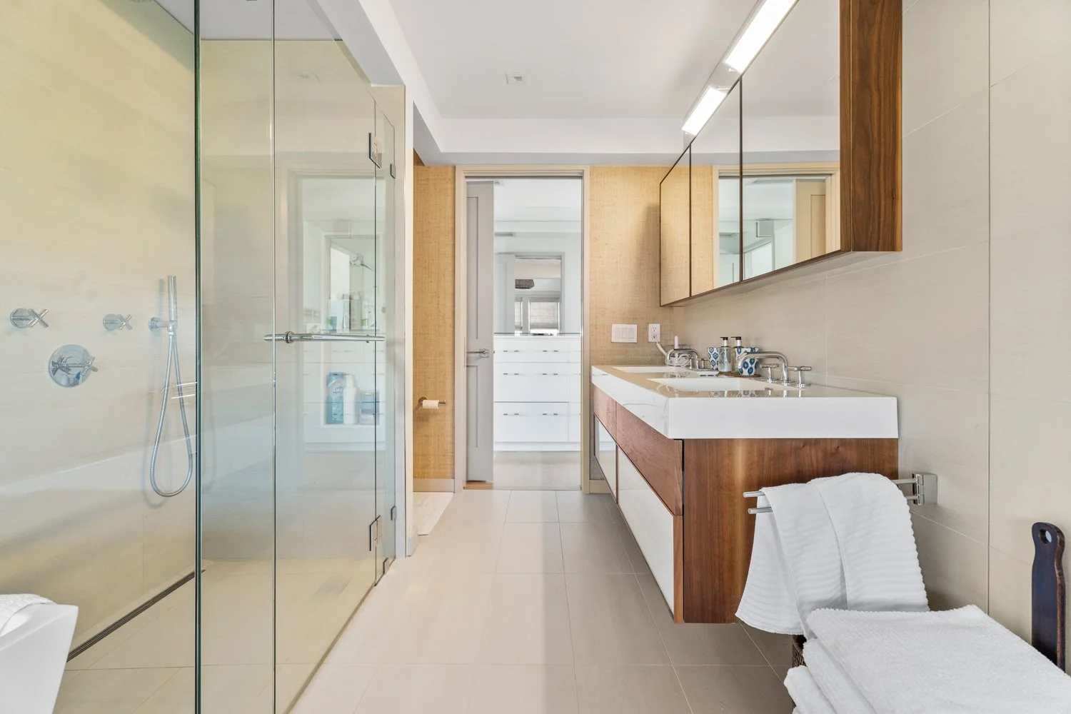 Modern bathroom with beige tiled walls and floor, glass shower enclosure, wooden vanity with double sinks, large mirror, and white towels on a bench.