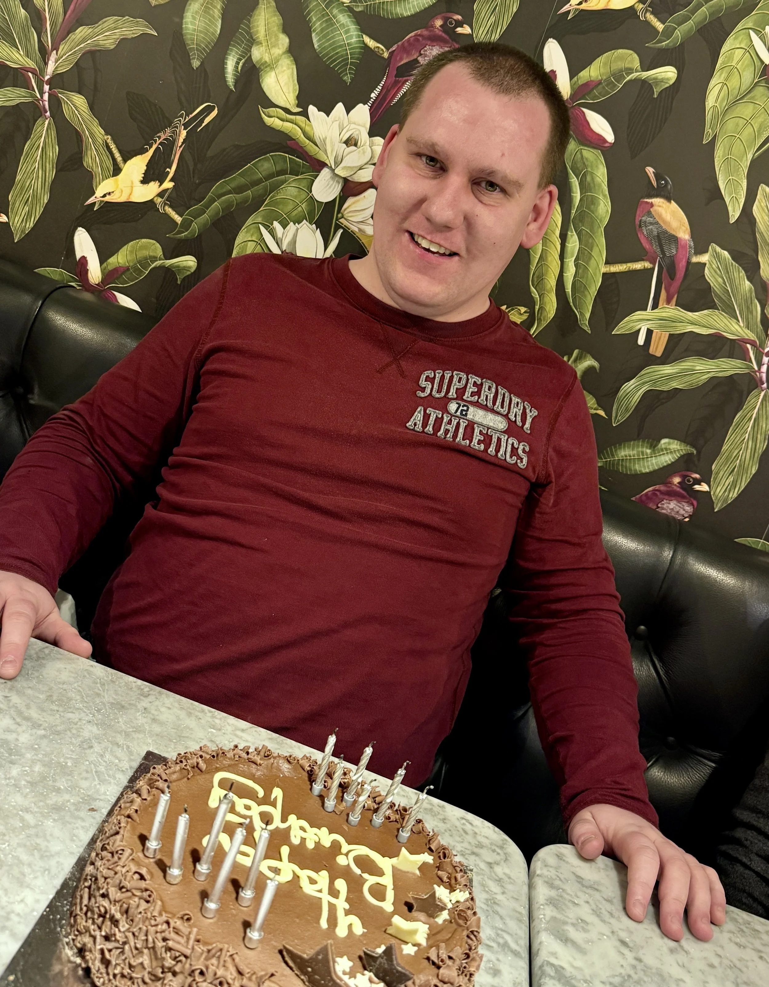 Person smiling with a chocolate birthday cake and candles, wearing a burgundy top.