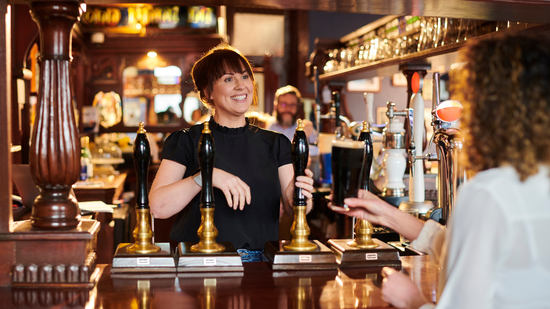 Person behind a pub bar smiling whilst serving a customer, with beer taps and bottles visible.