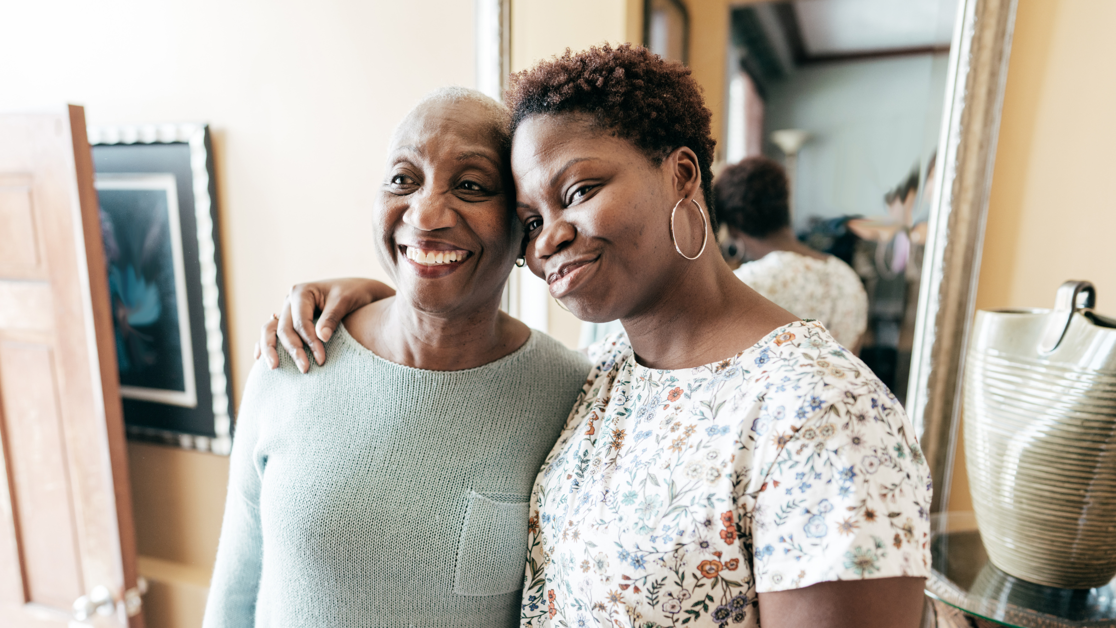 Two people with arms around each other smiling at camera in bright home setting, one wearing pale blue jumper, one wearing floral top with hoop earrings.