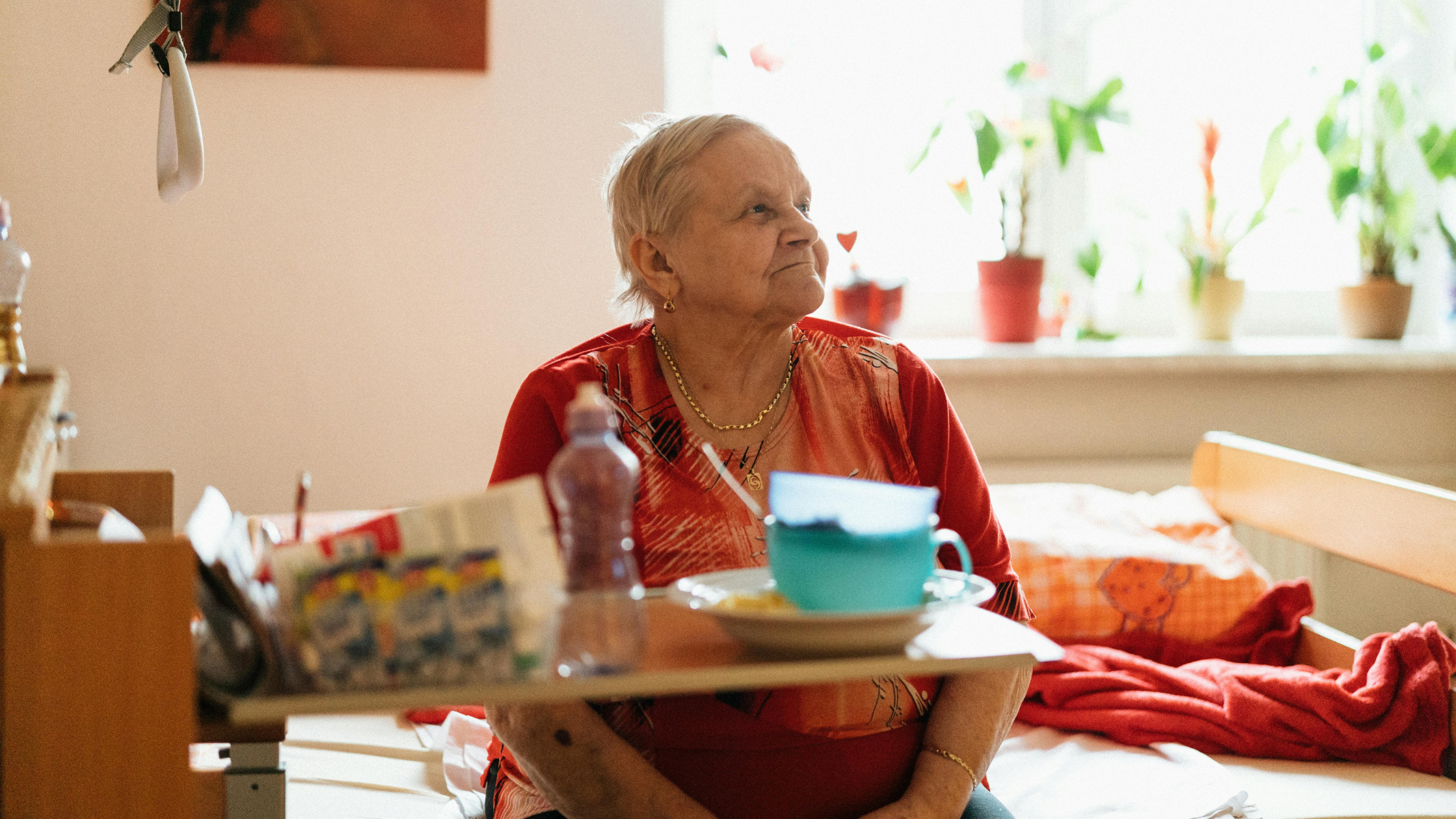 Person in orange top with gold necklace seated at table with turquoise mug and meal, bright room with plants on windowsill.