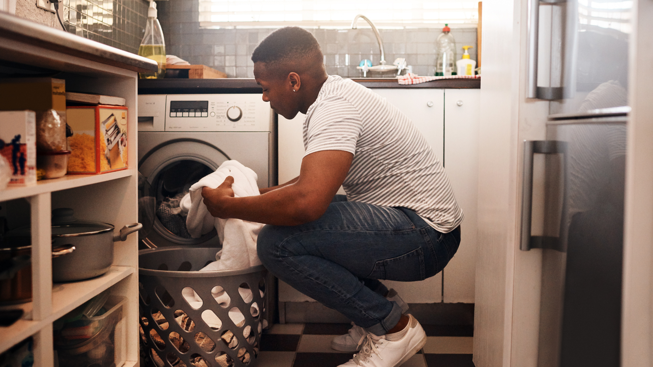 Person in striped top loading laundry from basket into washing machine in kitchen.