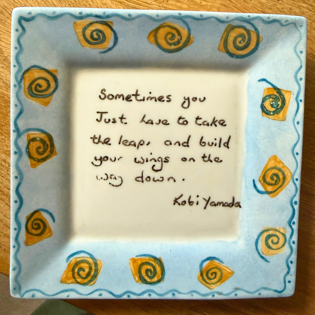 Ceramic square dish with blue and yellow spiral decoration. Text reads: "Sometimes you just have to take the leap, and build your wings on the way down. Koti Yamada"