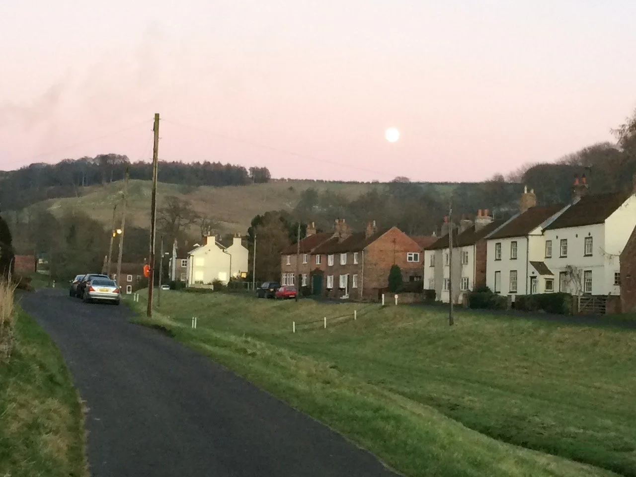 A quiet English village road at dusk, with a row of brick and white-rendered cottages, a green hillside, and a full moon visible in a pink sky.