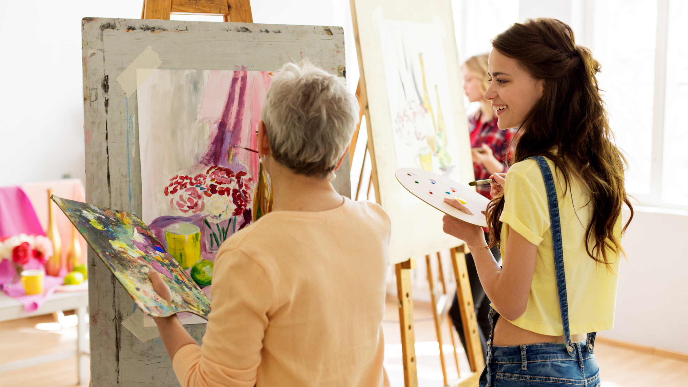 Two people in an art studio, one painting on a canvas whilst the other watches and smiles, holding a palette.