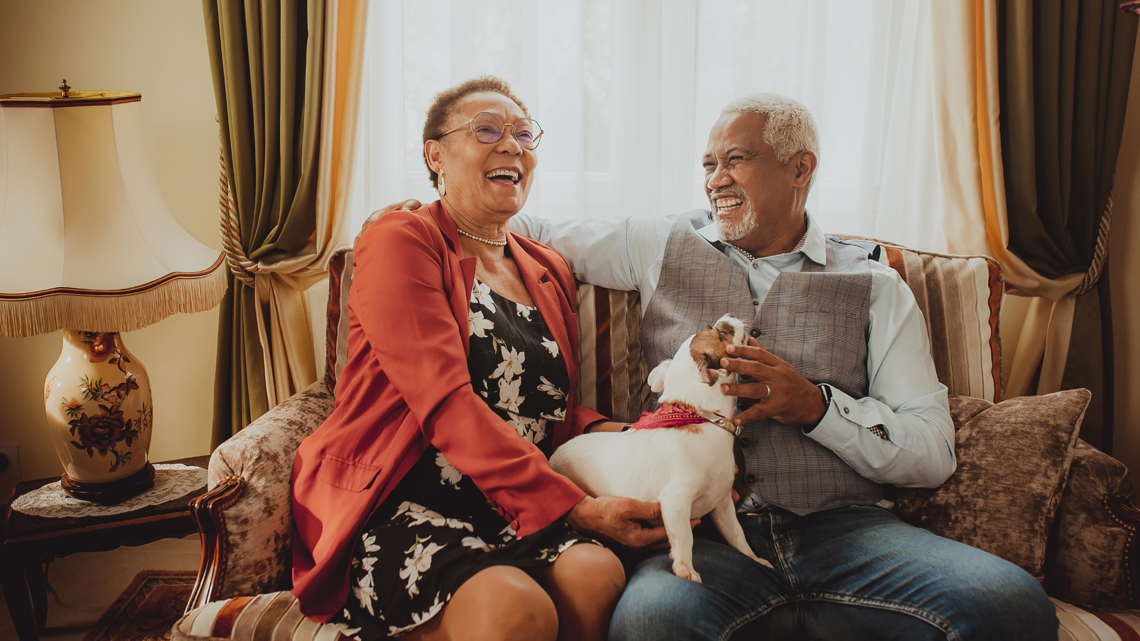 Two people sitting on a sofa laughing together, with a small white dog on their laps. Decorated living room with lamp, curtains and cushions in the background.