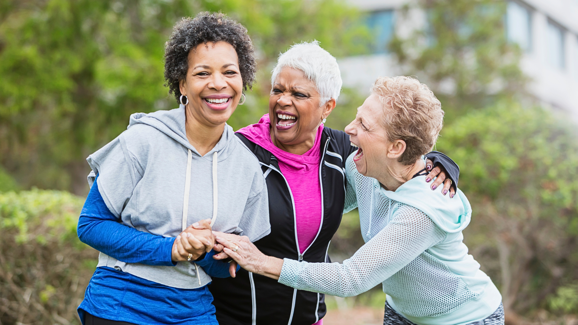Three people outdoors smiling together with arms linked, wearing sports clothing in a garden setting.