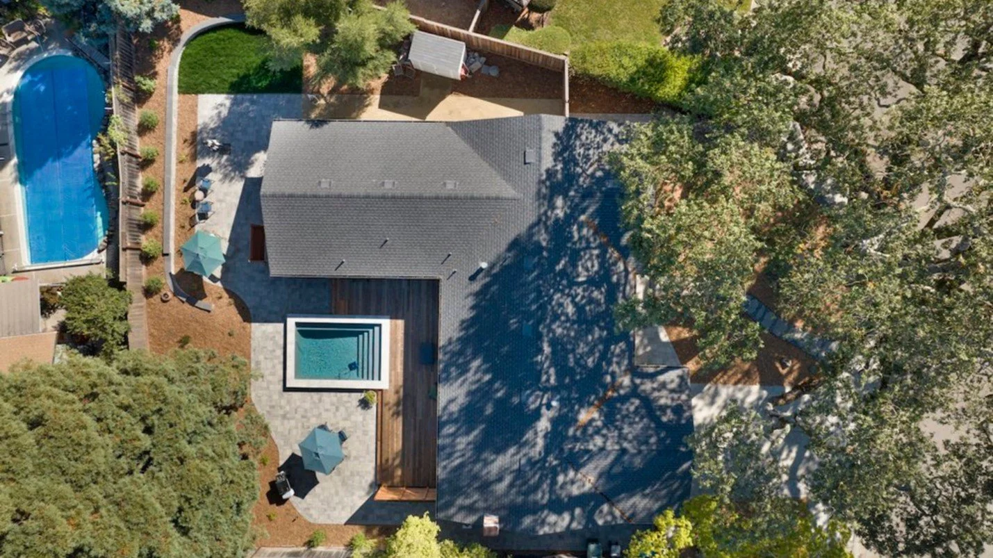 arial topo down view of a home with large trees in the front yard and a backyard featuring a pool with adjoining deck, decomposed granite patio seating area, and woodchipped landscape.