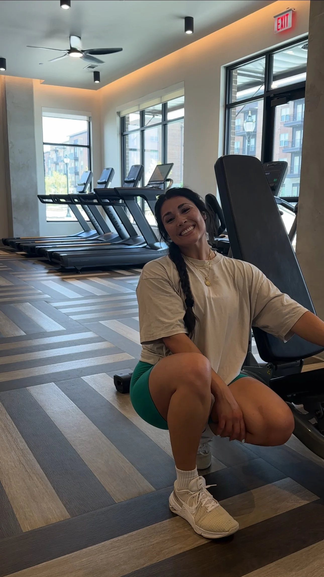 A woman smiling and squatting in a gym with treadmills in the background, large windows, and modern decor.