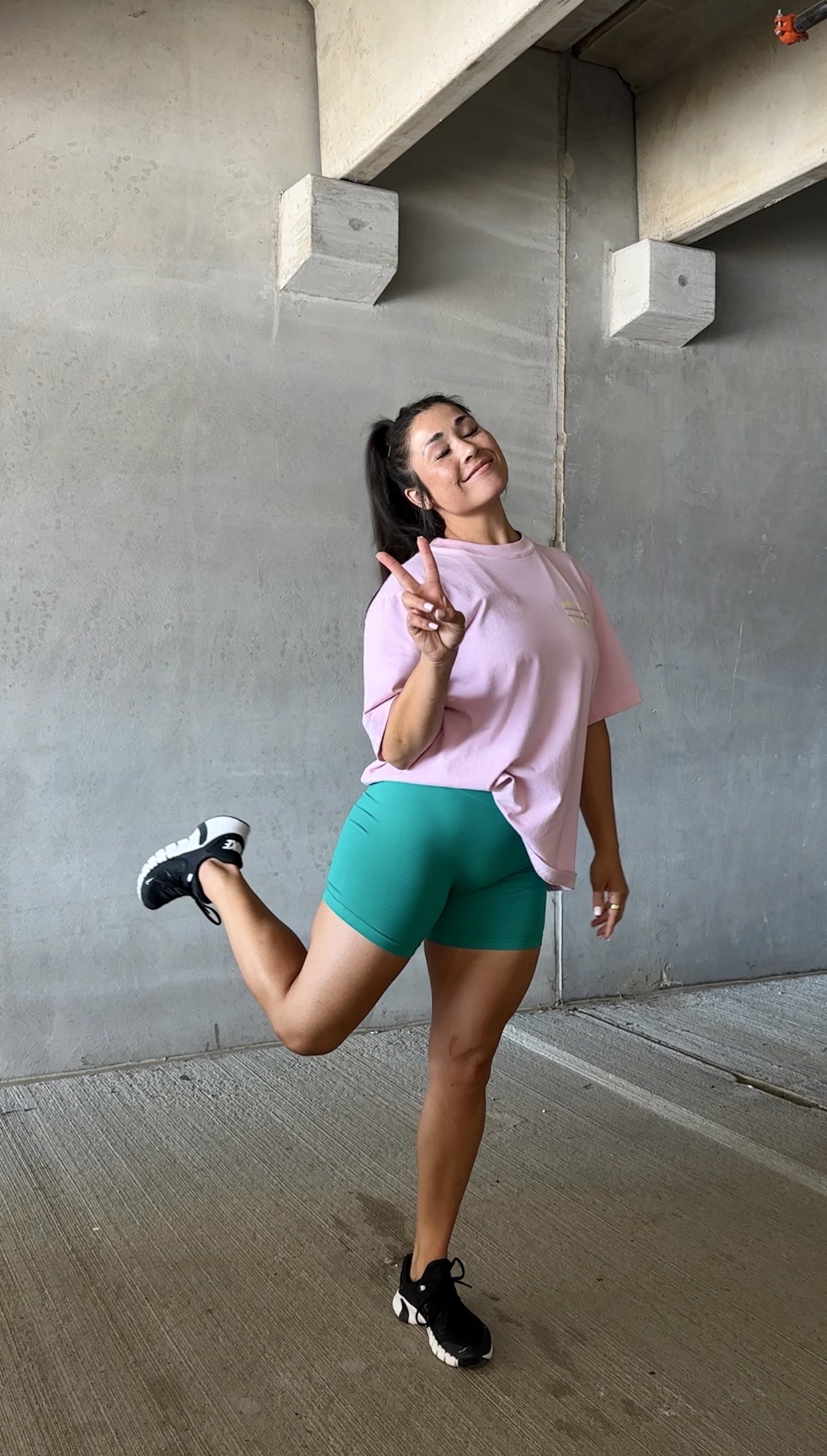 A woman in athletic clothing making a peace sign with her right hand, standing on one leg with her left leg bent upward behind her, smiling with eyes closed, in front of a concrete wall.