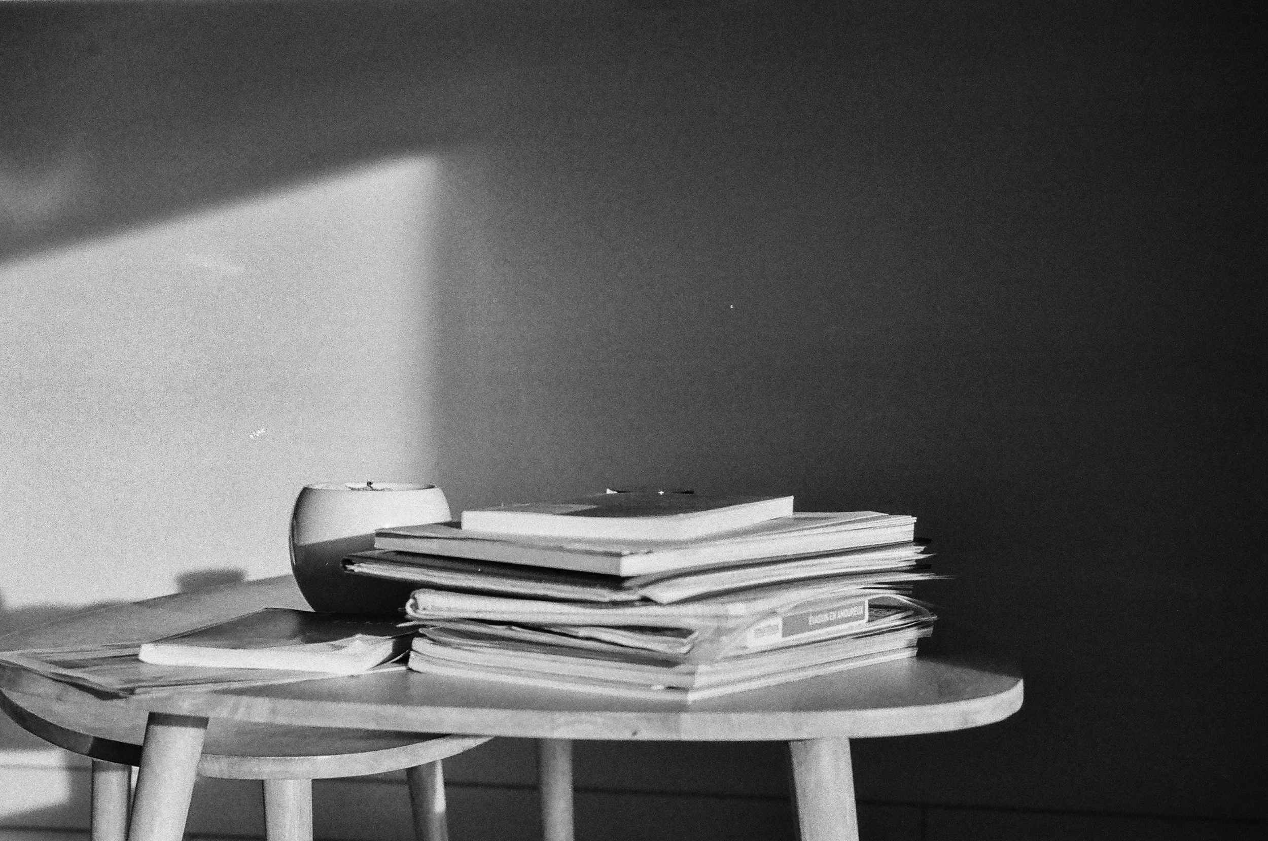 A stack of folders and magazines on a small round wooden table, with a cup or mug and some paper or notebooks nearby, in a dimly lit room.