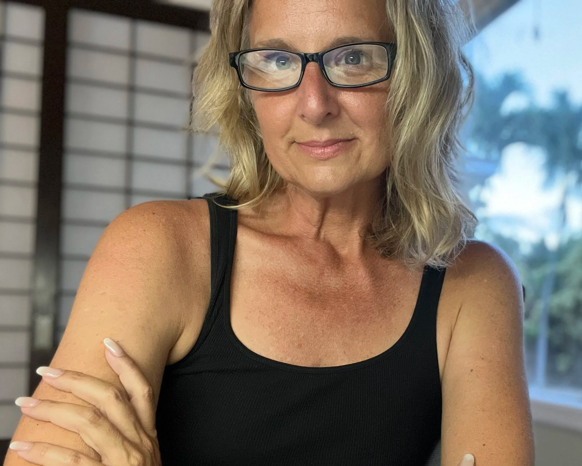 Close-up of a woman with blonde hair wearing glasses, dressed in a black tank top, sitting with her arms crossed.