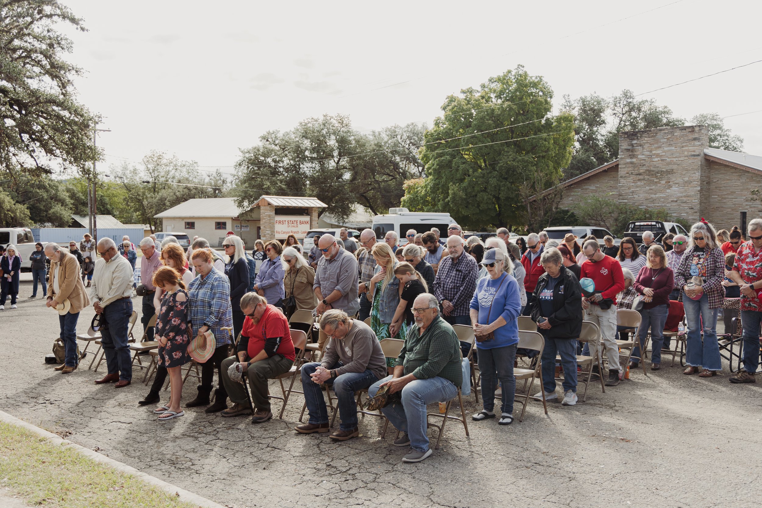 A large group of people praying outdoors, several chairs set up, on a cracked asphalt surface with trees and buildings in the background.