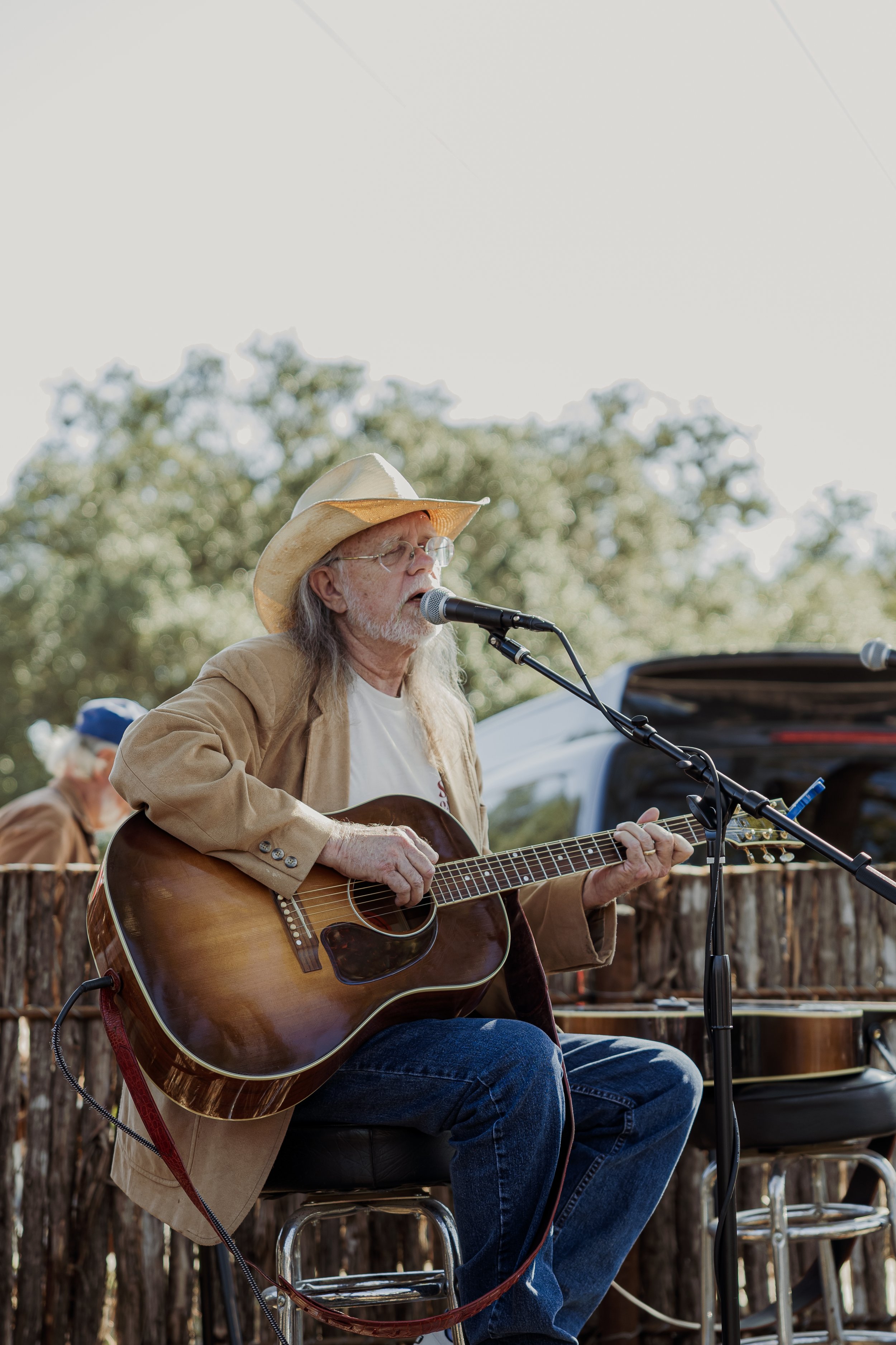 An elderly man wearing a cowboy hat and glasses, sitting on a stool, playing an acoustic guitar and singing into a microphone outdoors during daytime.