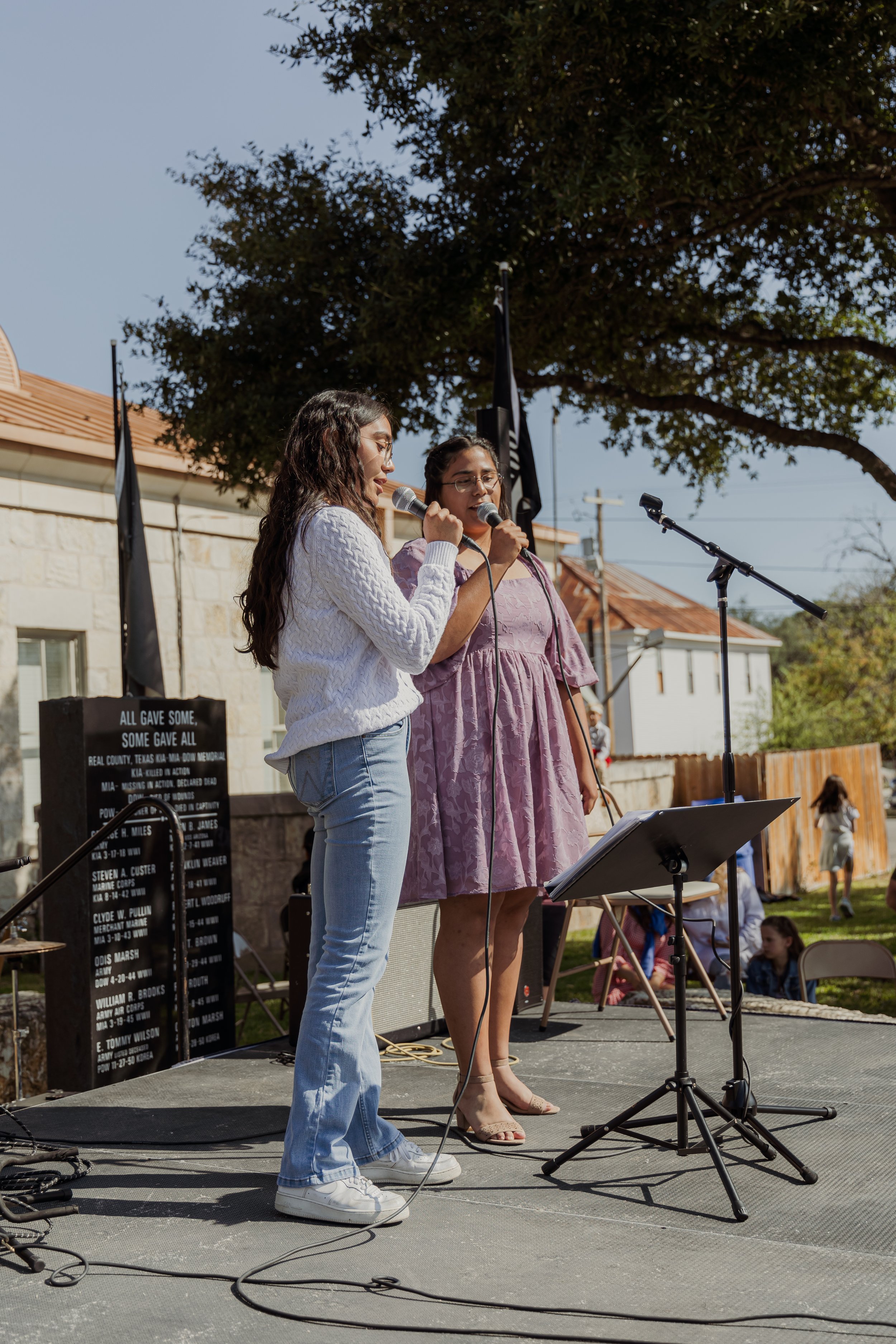 Two women singing into microphones on an outdoor stage, with a notice board and a large tree in the background.