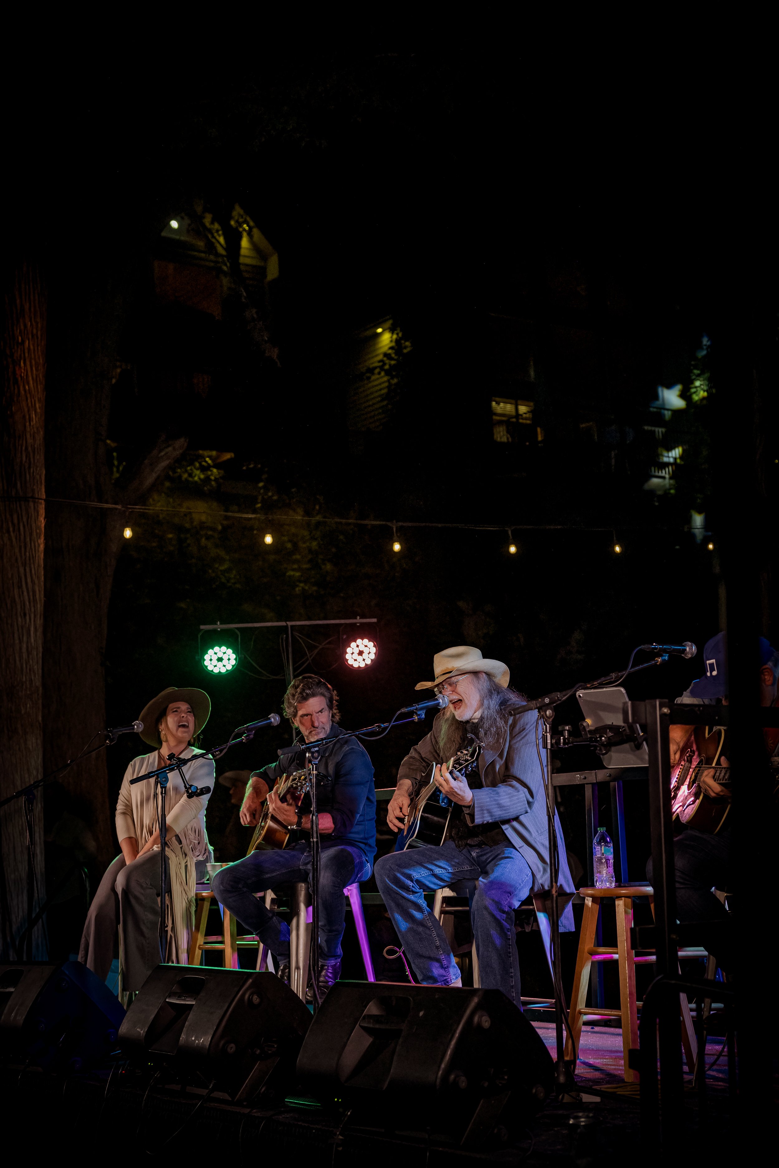 Four musicians performing on an outdoor stage at night, playing guitars and singing into microphones, with colorful stage lights and string lights overhead.