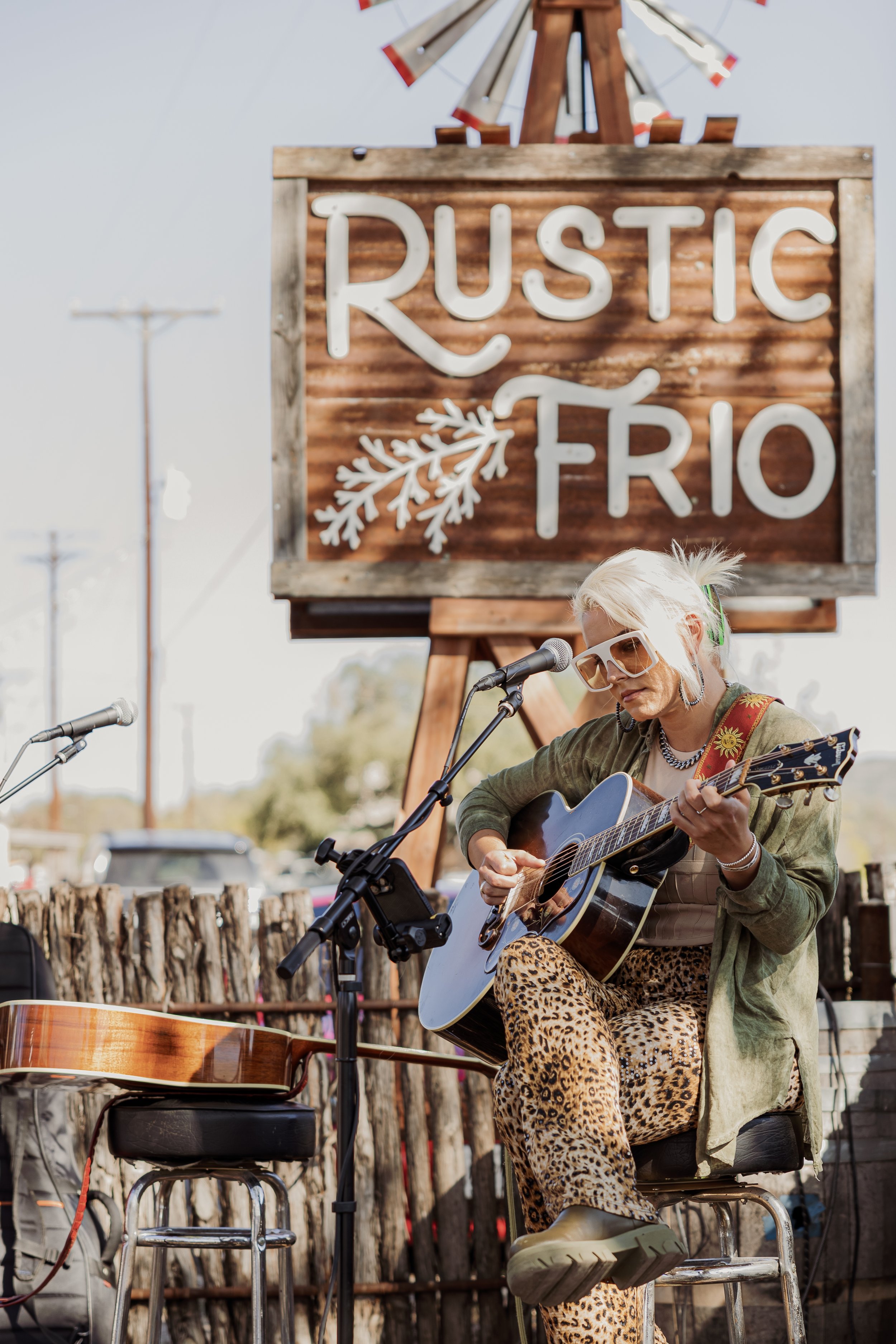 A woman with platinum blonde hair, wearing sunglasses, playing an acoustic guitar at an outdoor event with a rustic sign that reads 'Rustic Rio' and a windmill in the background.