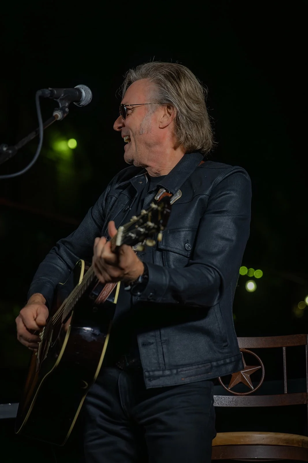 A man wearing sunglasses and a denim jacket playing an acoustic guitar and singing into a microphone.
