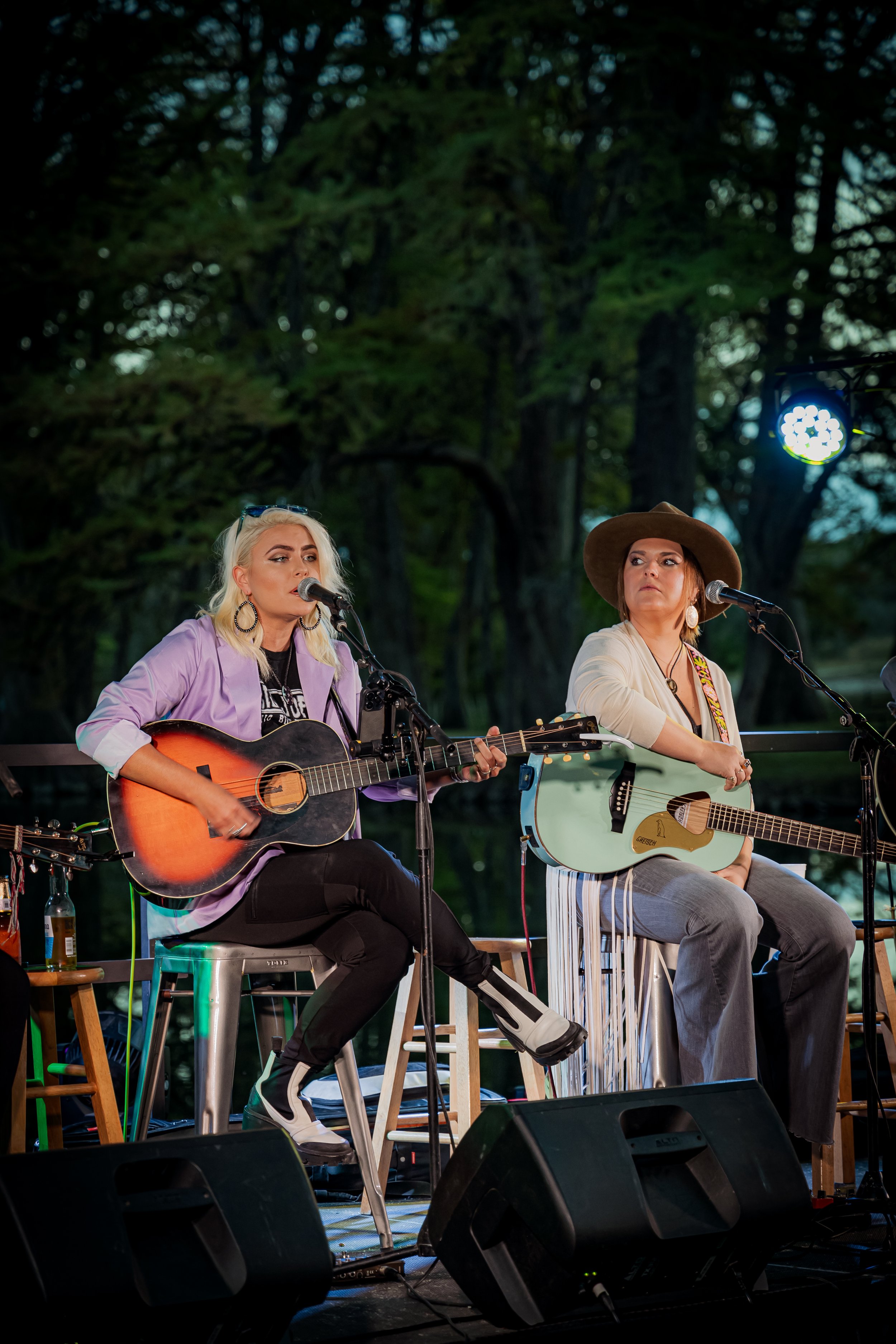 Two women performing on an outdoor stage, playing acoustic guitars, with microphones, in a forested area.