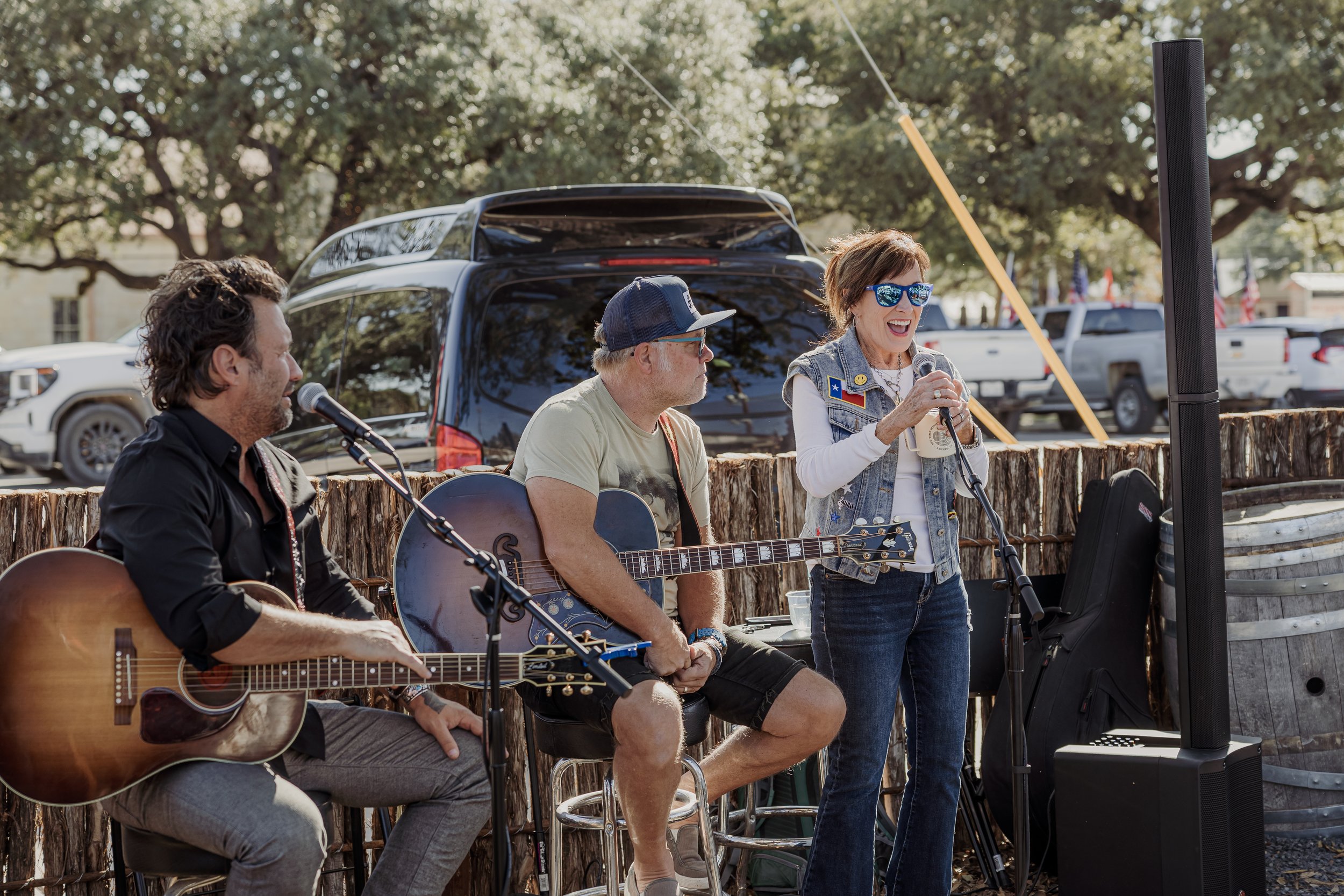 Three musicians performing outdoors, two men playing guitars and a woman singing into a microphone, in front of a wooden fence with parked cars and trees in the background.