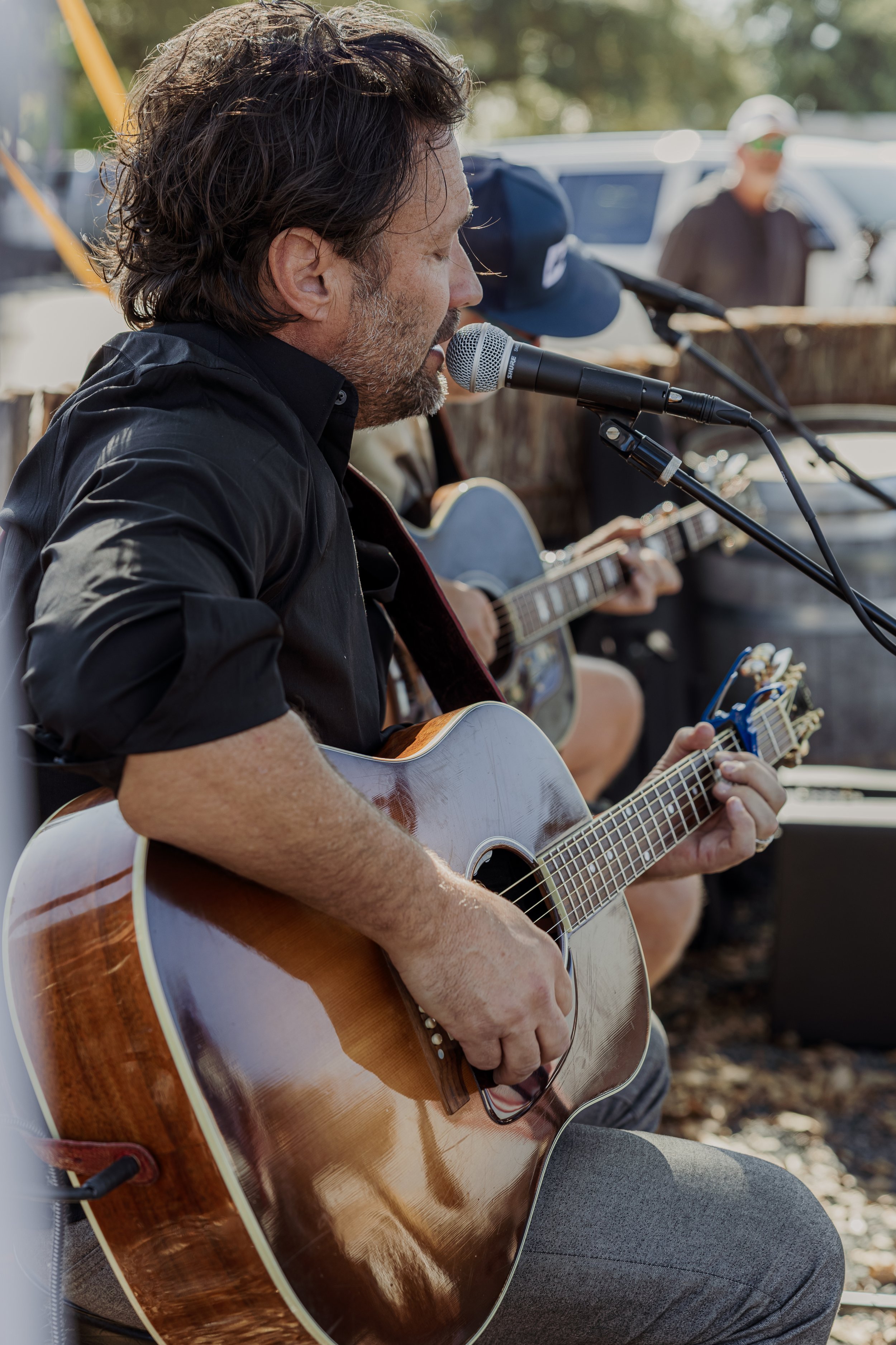 A man with dark hair and a beard playing an acoustic guitar and singing into a microphone outdoors, with another person in the background also playing guitar.