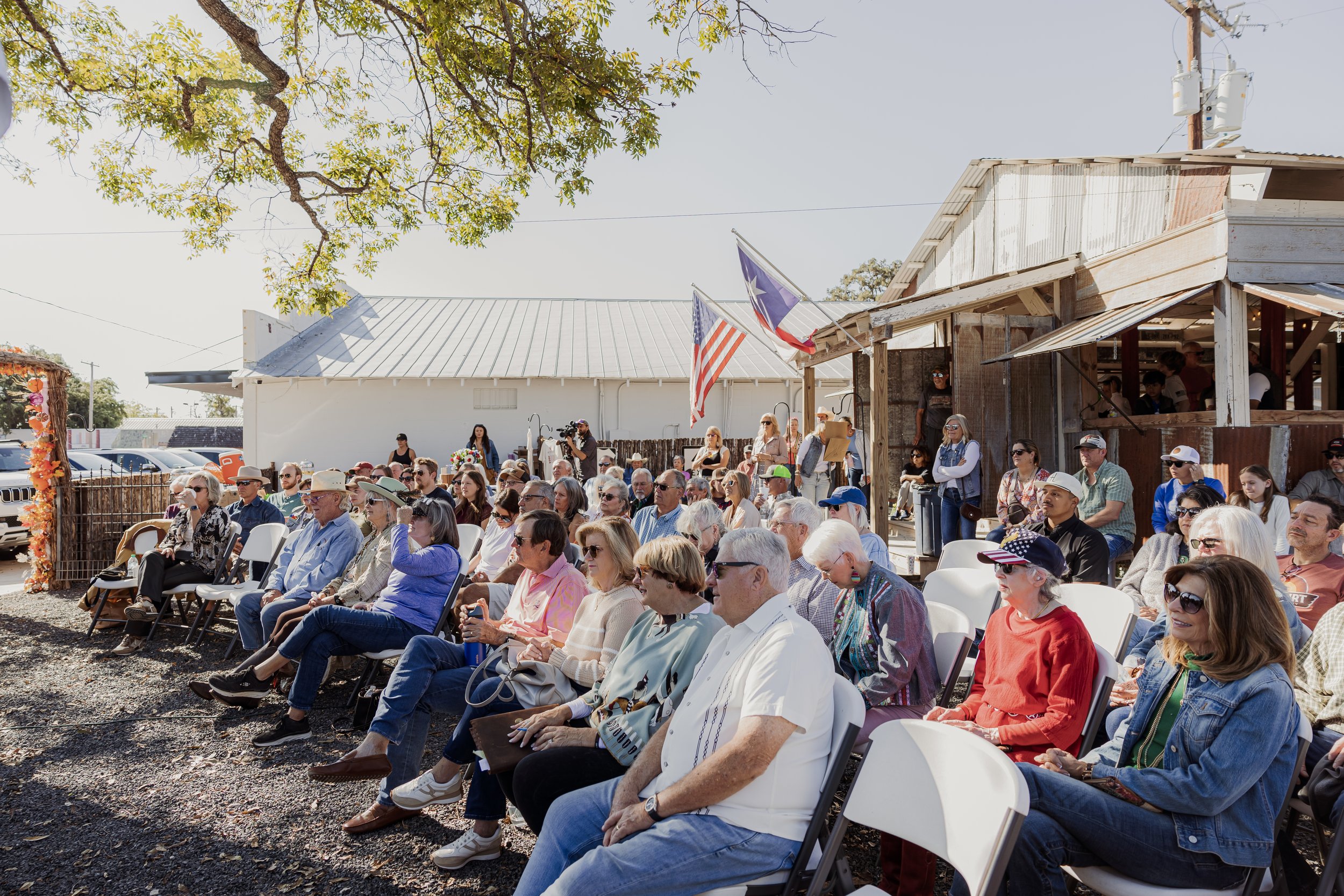 An outdoor event with a large group of people seated on chairs, attending a gathering or ceremony in front of a rustic building in Texas, adorned with American and Texas flags.
