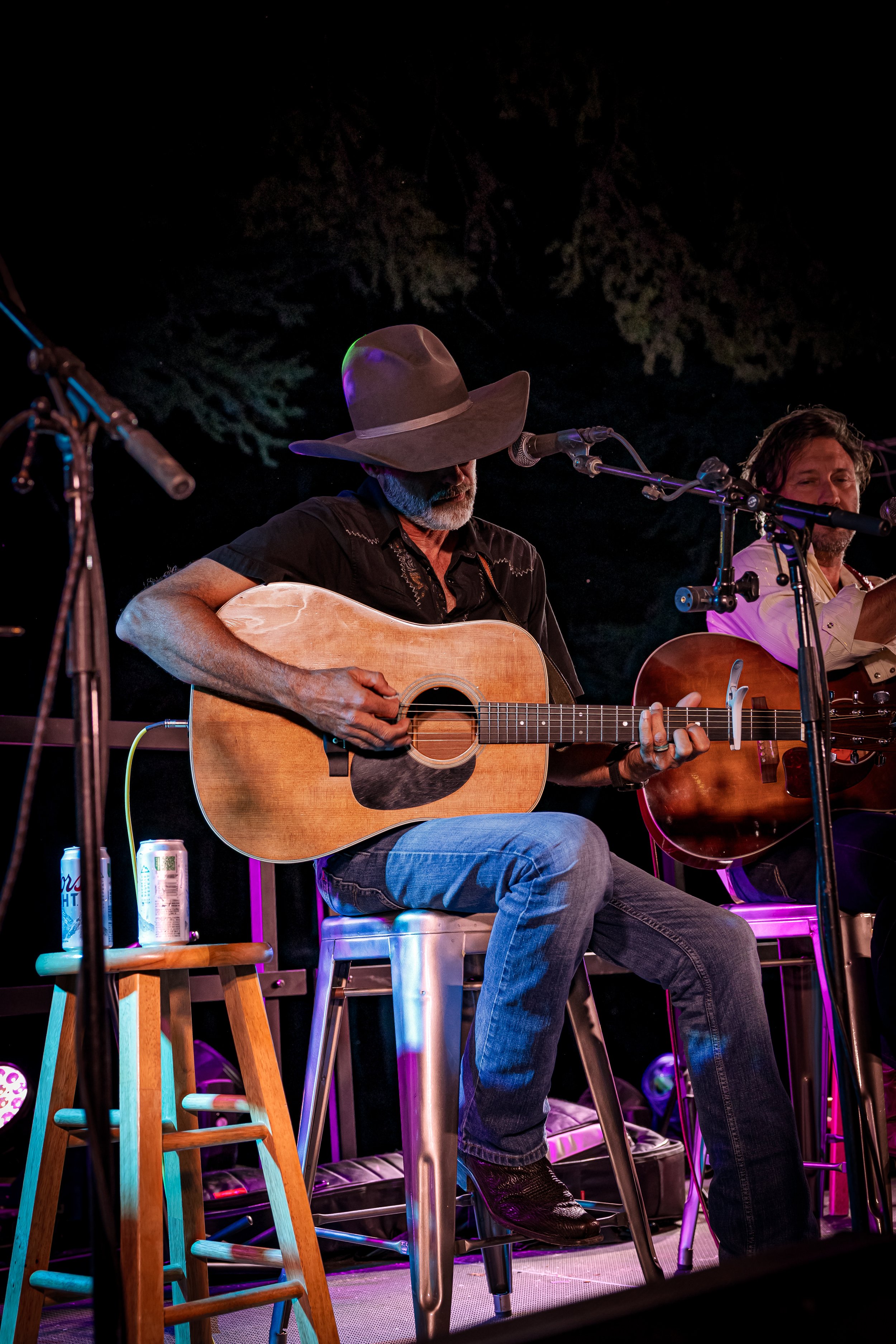 A man wearing a hat plays an acoustic guitar during a live music performance on stage, with another musician playing guitar beside him.