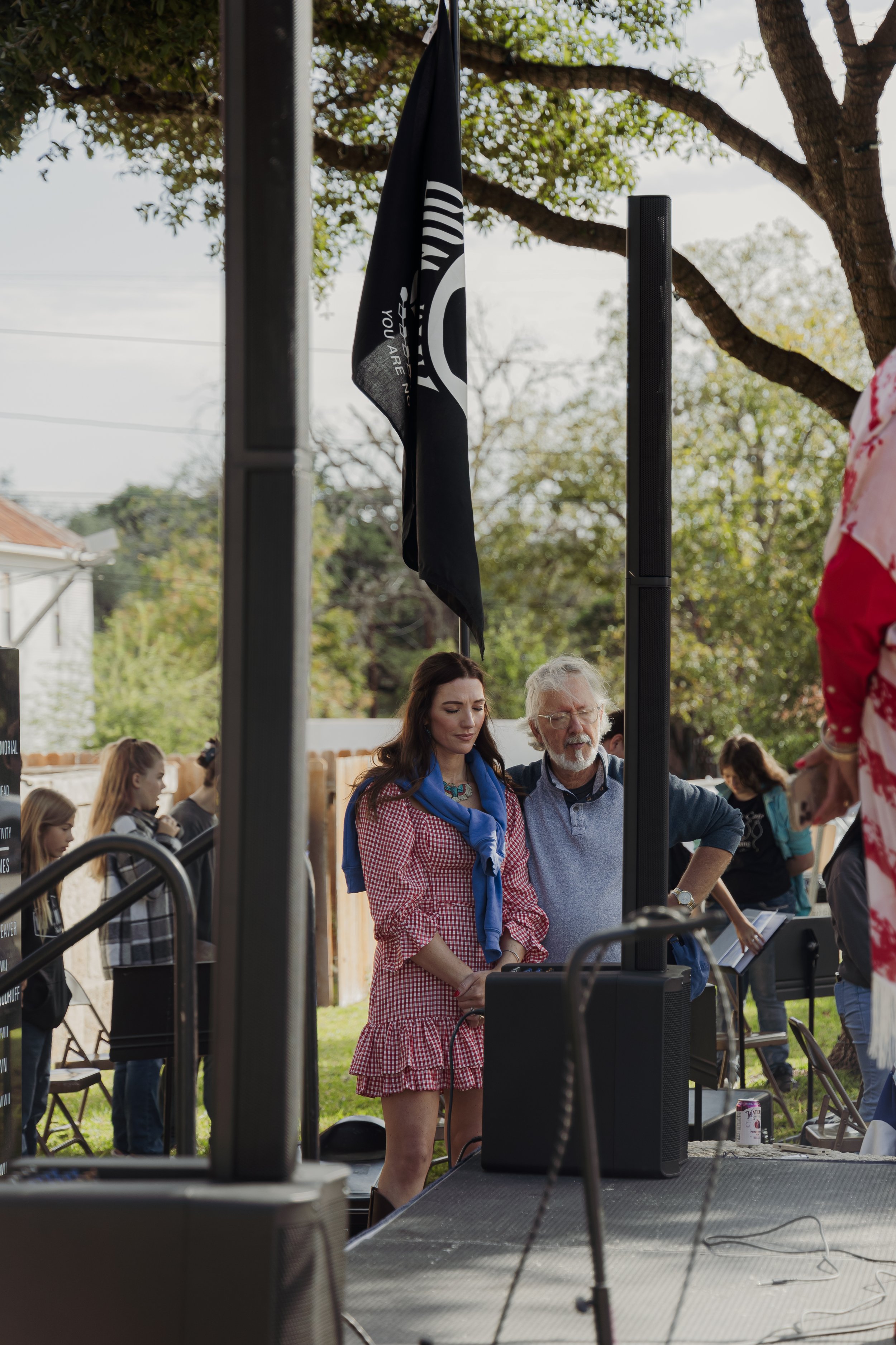 Two people standing outdoors near a speaker and flag, with trees and other people in the background.