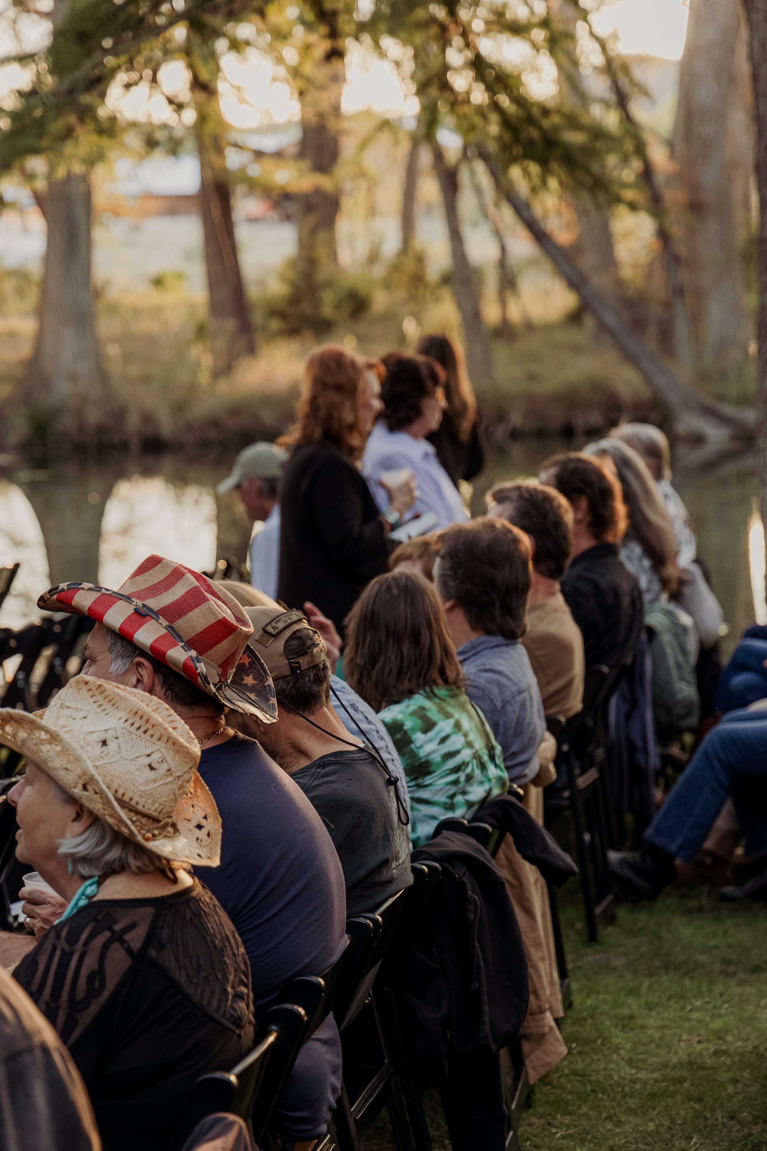 People seated outdoors near a lake during sunset for an event, with some women standing and speaking.