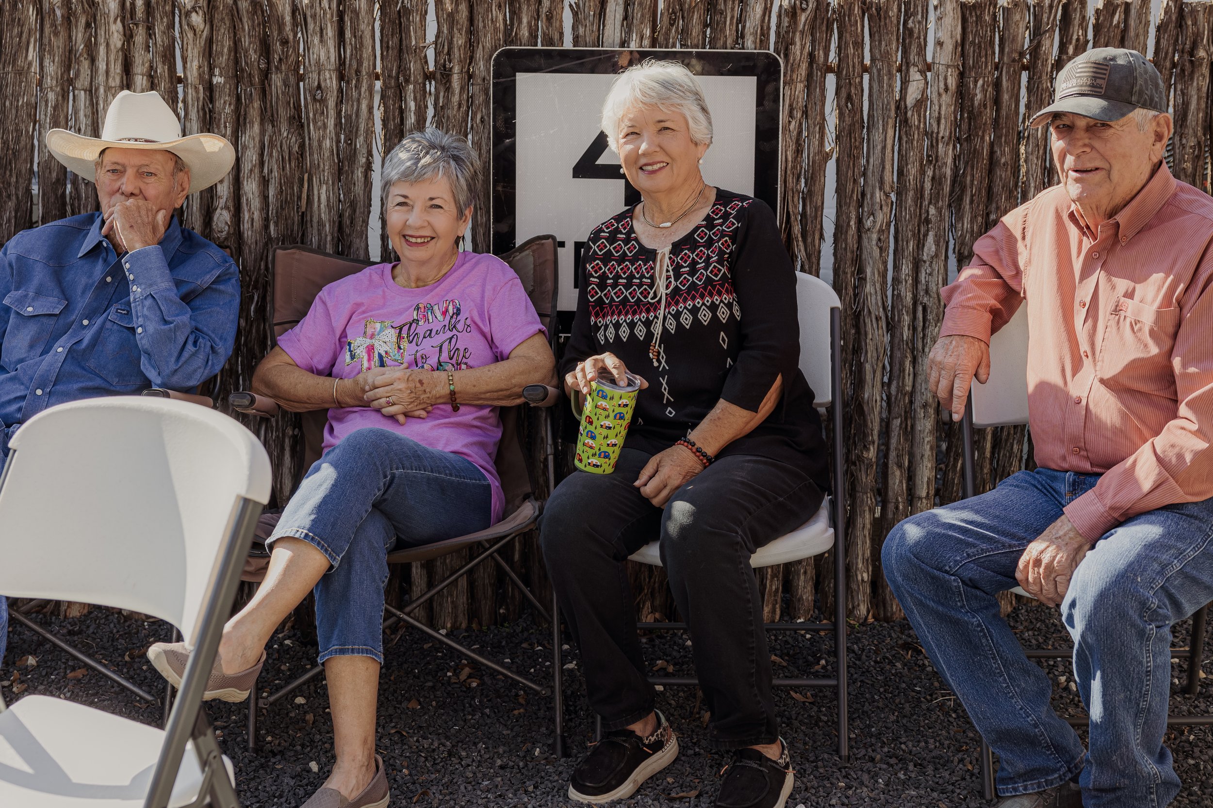Four elderly people sitting outdoors in front of a wooden fence, smiling and talking, with folding chairs around them.