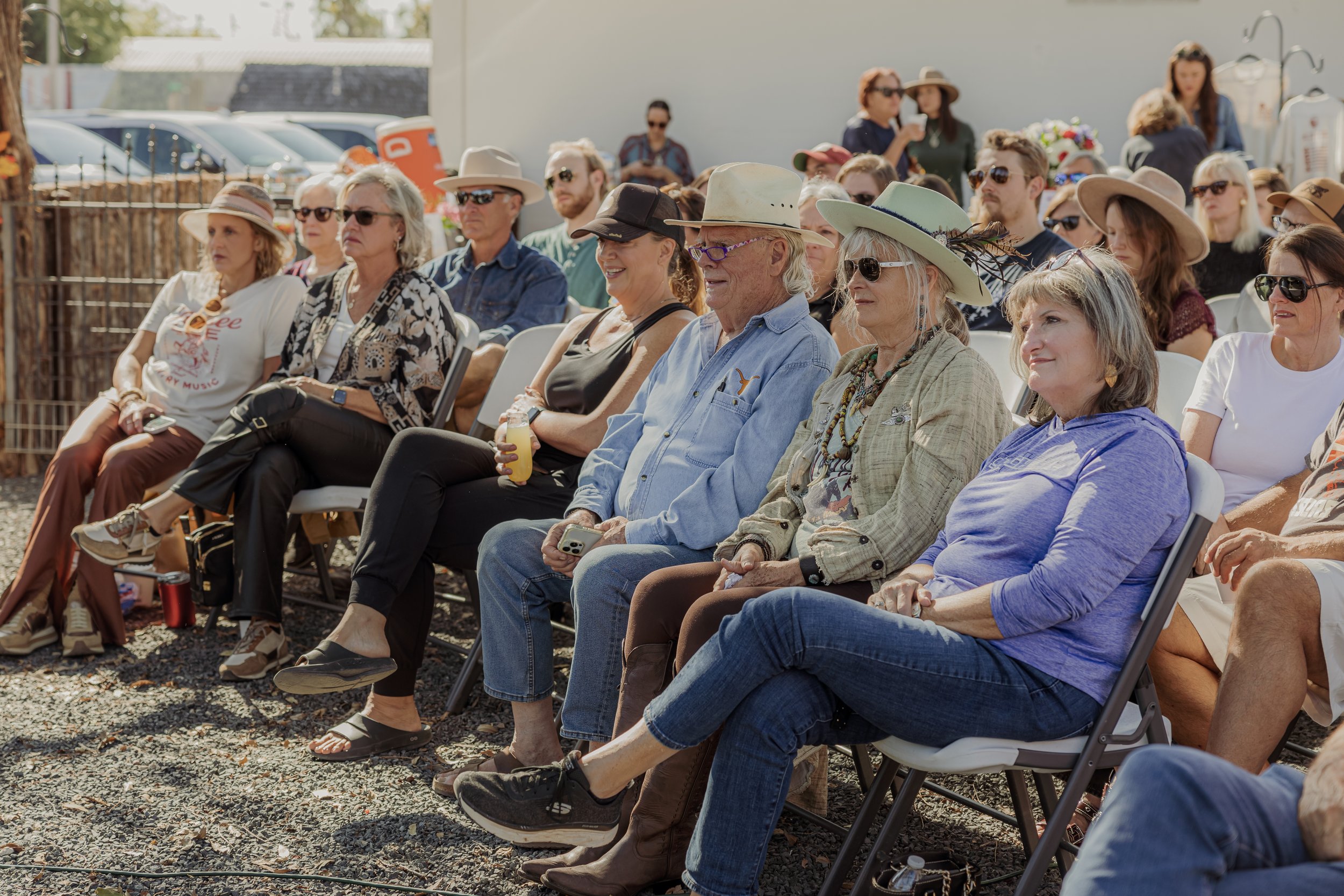 A group of people sitting outdoors on folding chairs at an event, wearing casual clothing and sun hats, with some holding drinks and phones.