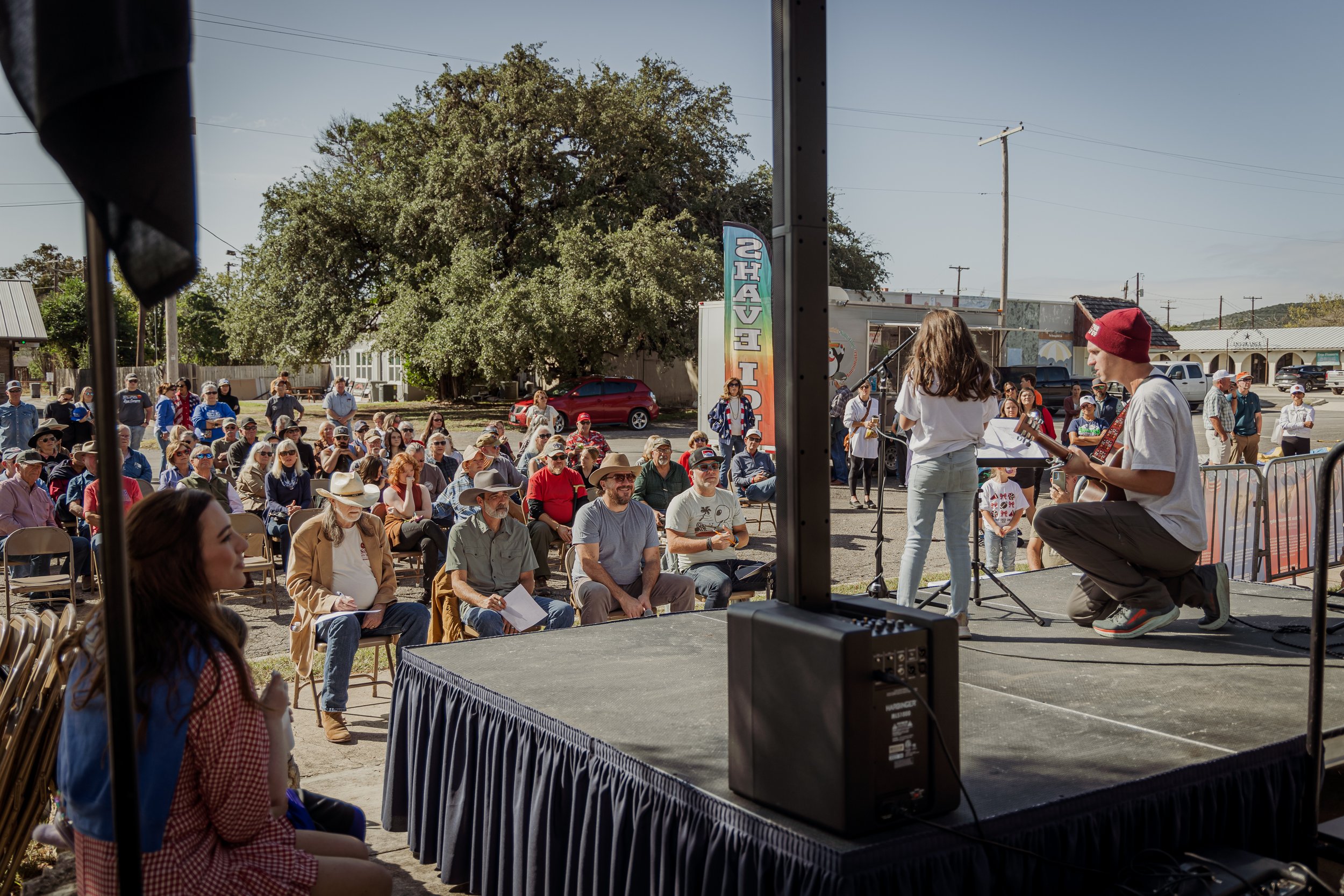 Outdoor event with people seated on chairs watching a performance on a small stage, featuring a girl singing and a young man playing guitar, under a large tree on a sunny day in a small town