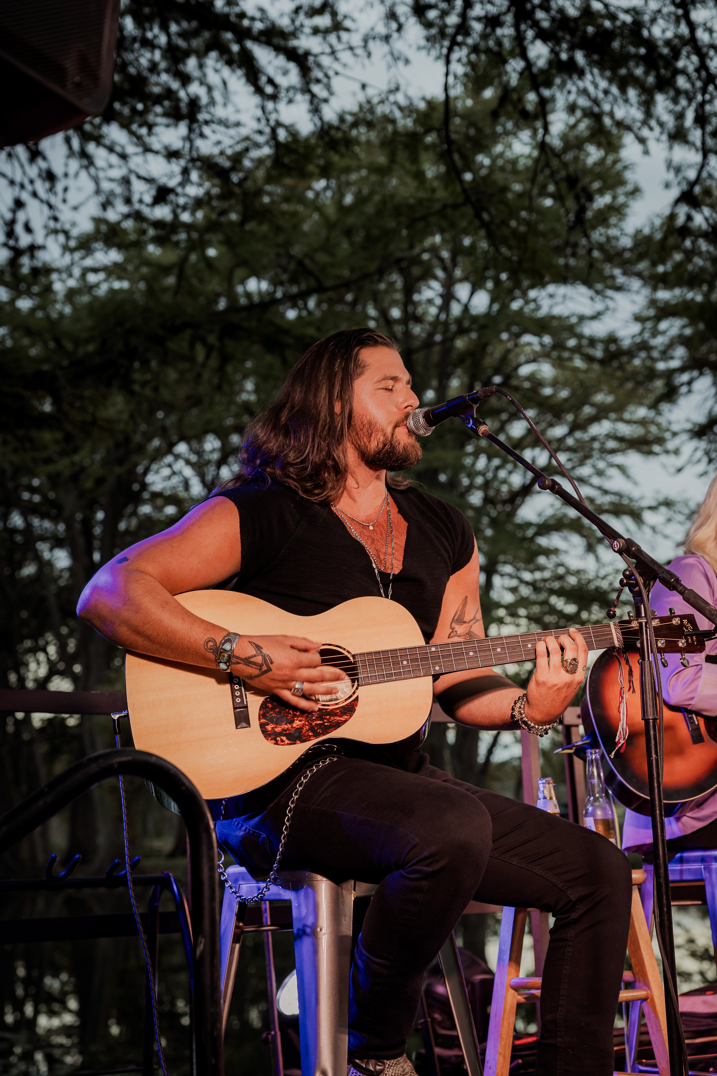 Man with long hair and tattoos playing acoustic guitar and singing into a microphone outdoors during sunset.