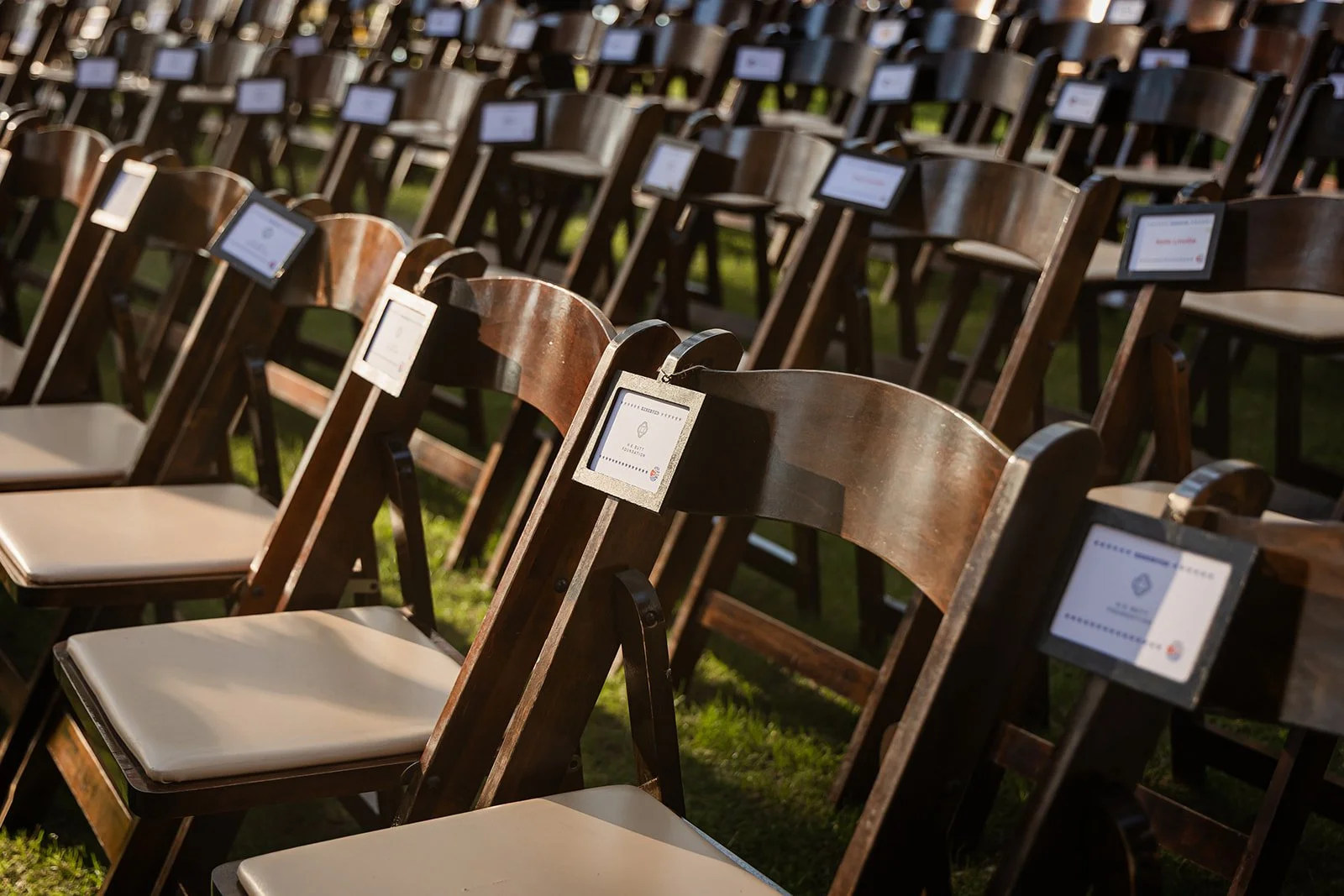 Multiple wooden chairs with white cushions arranged in rows outdoors, each with a small screen attached to the back of the seat in natural sunlight.