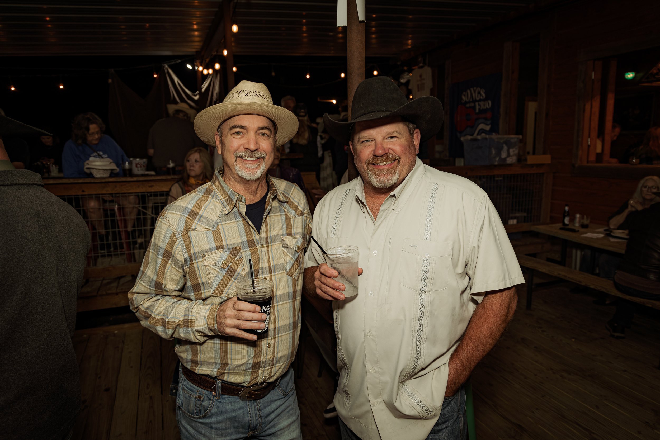Two middle-aged men with gray beards and cowboy hats smiling and holding drinks at a social gathering in a rustic wooden venue.