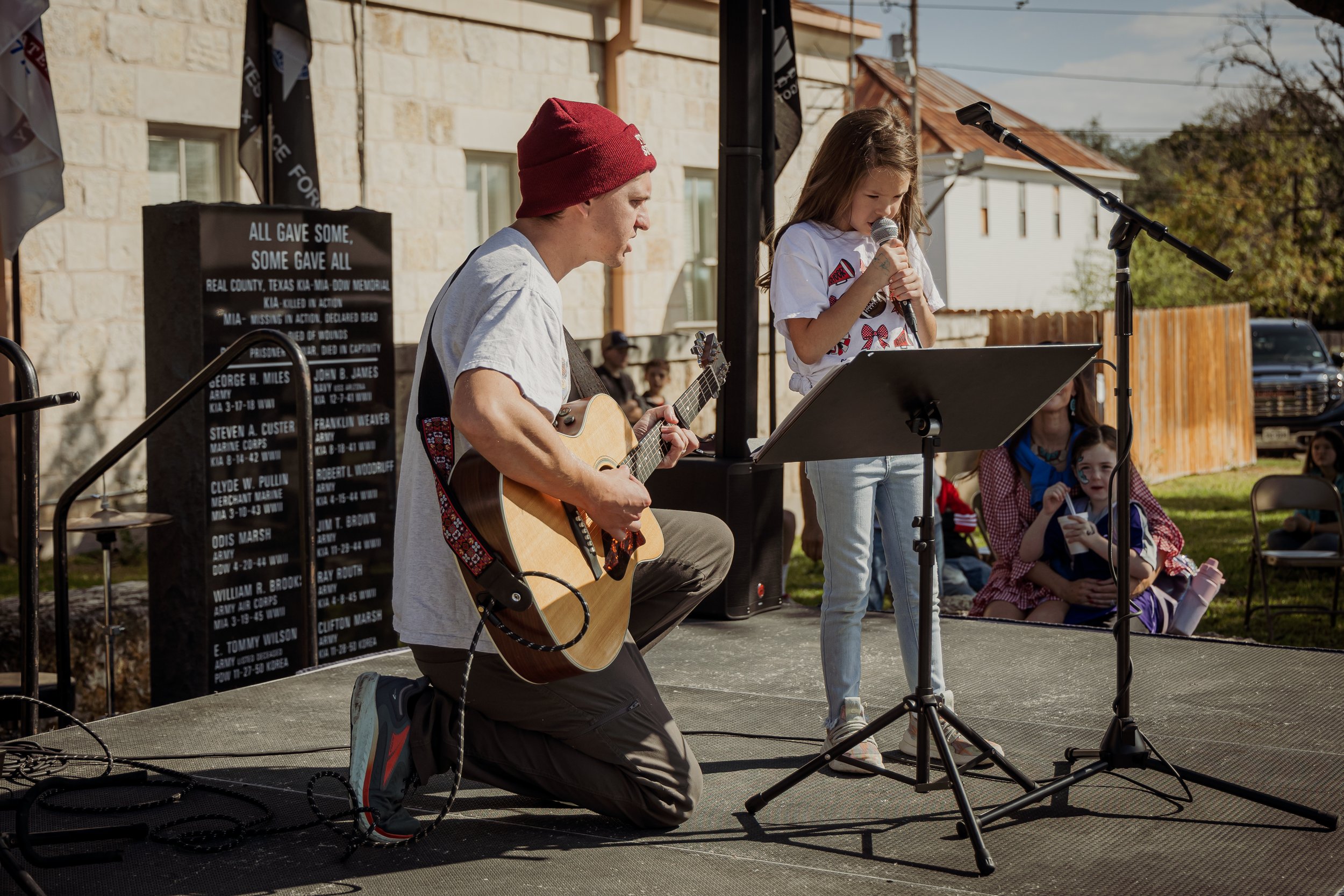 A man kneeling and playing guitar, a girl standing and singing into a microphone, on an outdoor stage with a small audience in daytime.