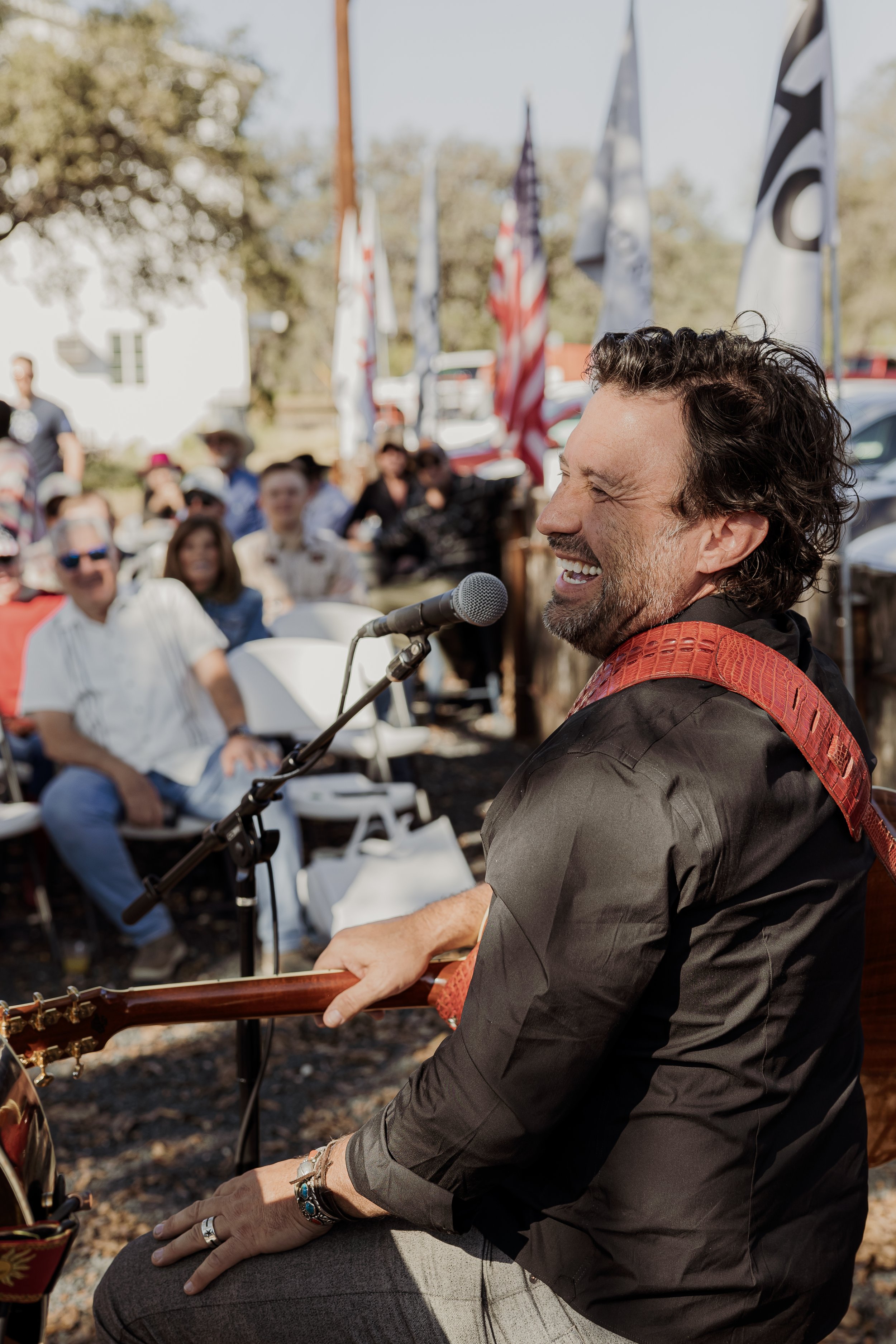 A man with curly hair and a beard playing an acoustic guitar and singing into a microphone at an outdoor event with people seated in front of him and American flags and flags in the background.