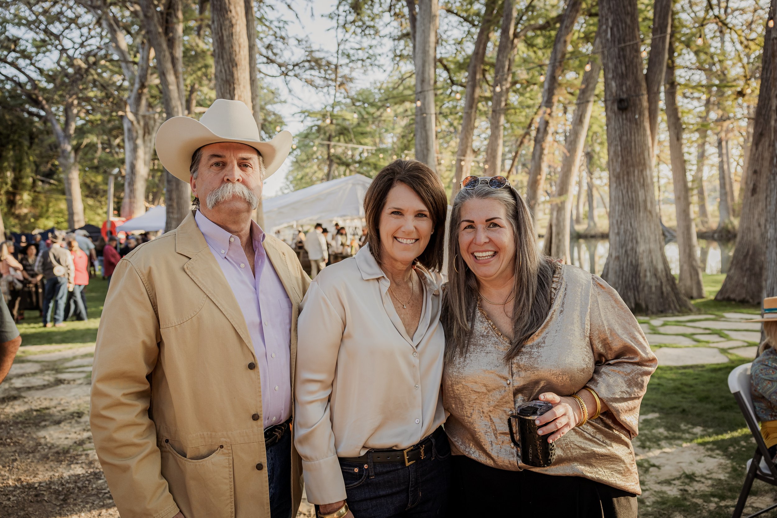 Three smiling women and a man at an outdoor event with trees and tents in the background