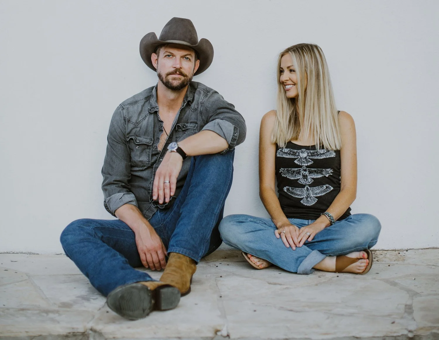 A man wearing a cowboy hat and denim jacket sitting cross-legged with a woman in a black shirt with bird patterns and jeans sitting against a white wall.