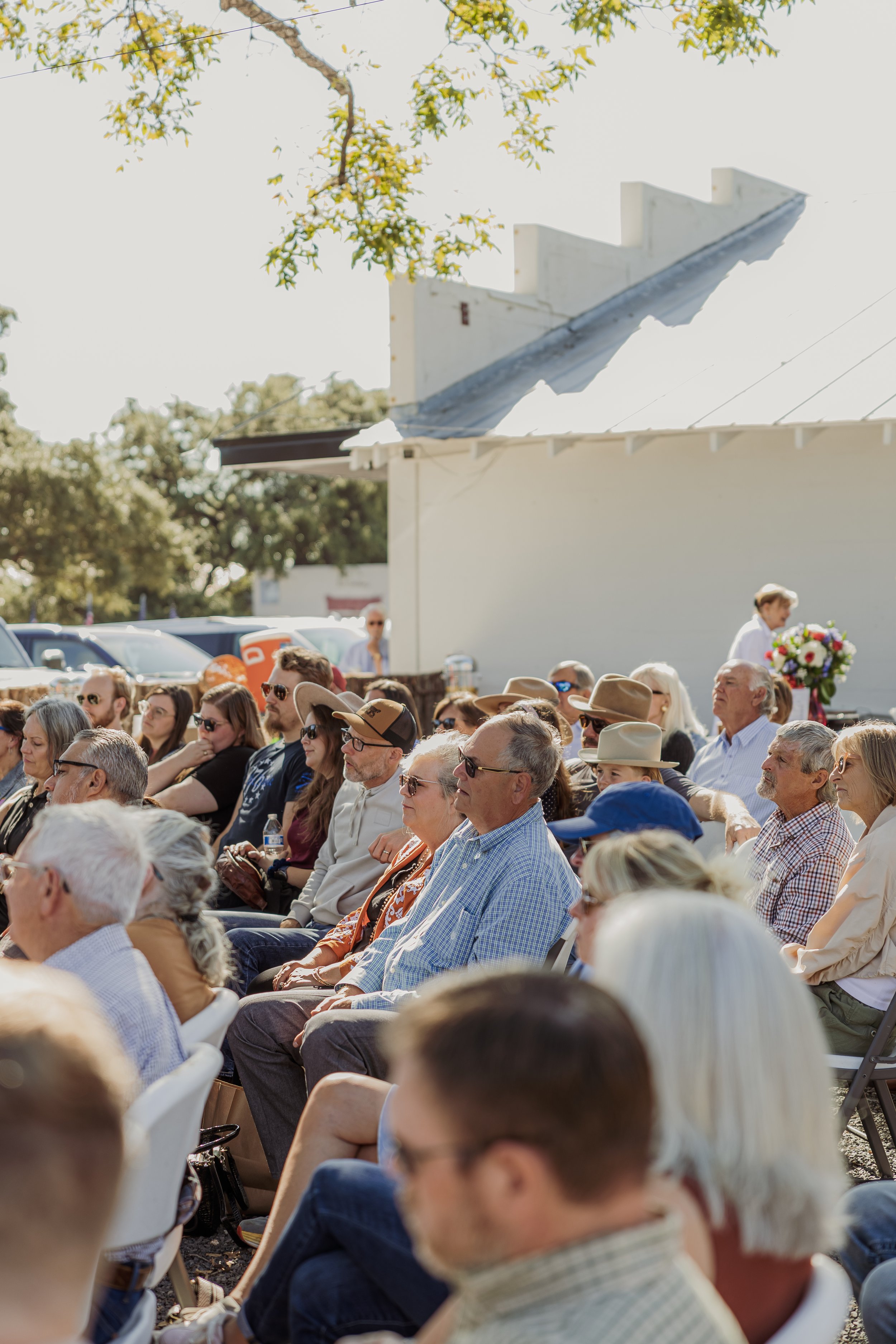 An outdoor crowd sitting in chairs on a sunny day, with some wearing hats and sunglasses, during what appears to be a community event or gathering.