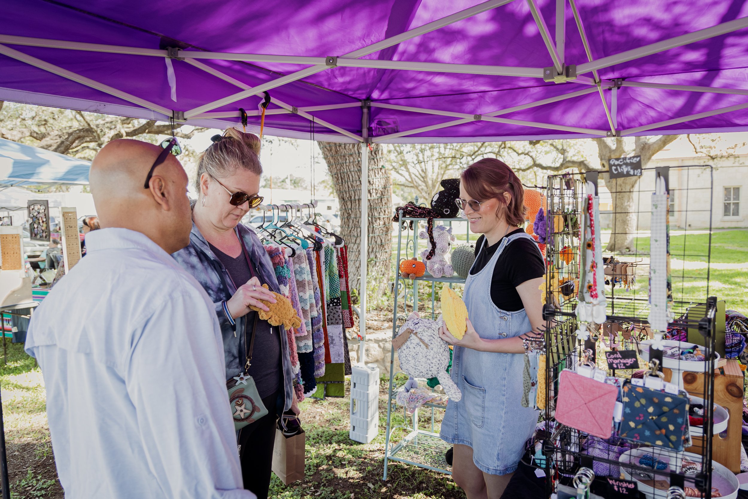 A vendor at a craft fair stall sells knitted and fabric accessories to a woman and a man, under a purple canopy with trees in the background.