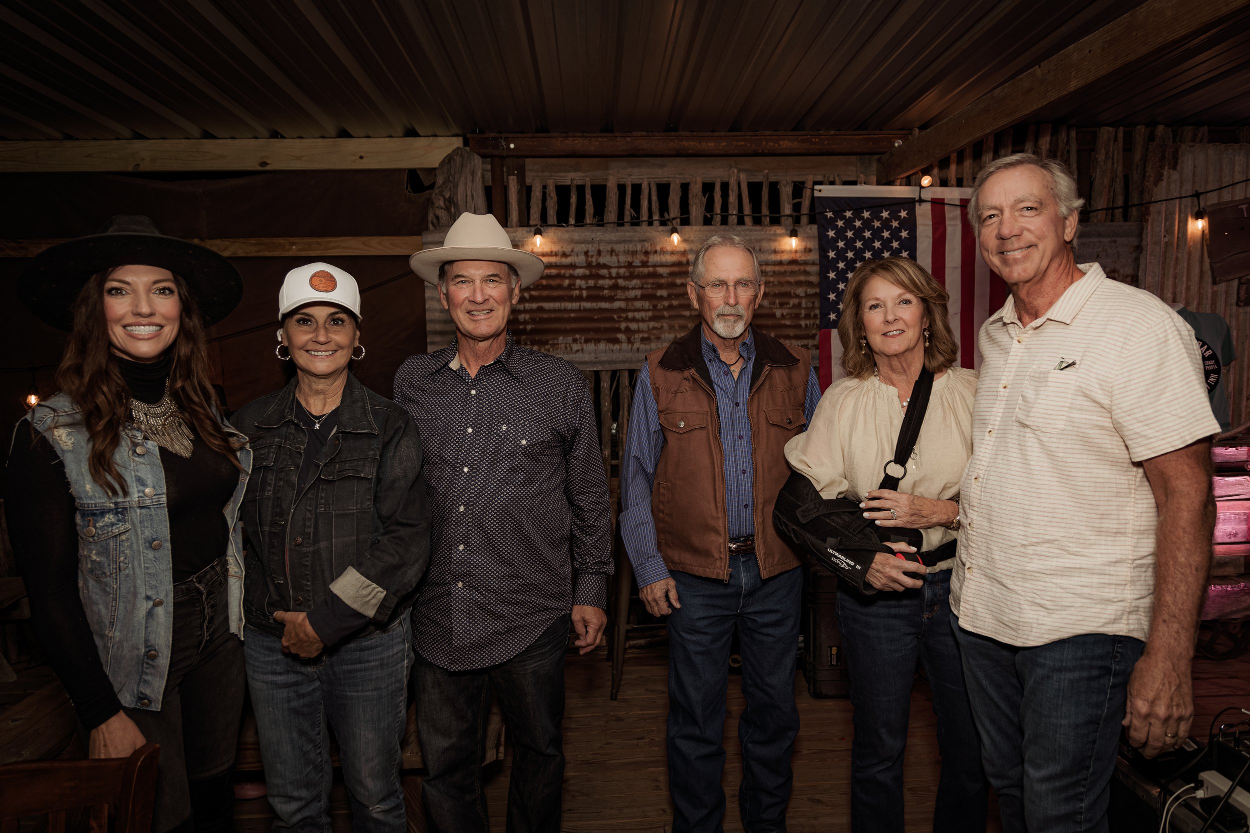 Group of seven people posing together indoors with American flag in background.