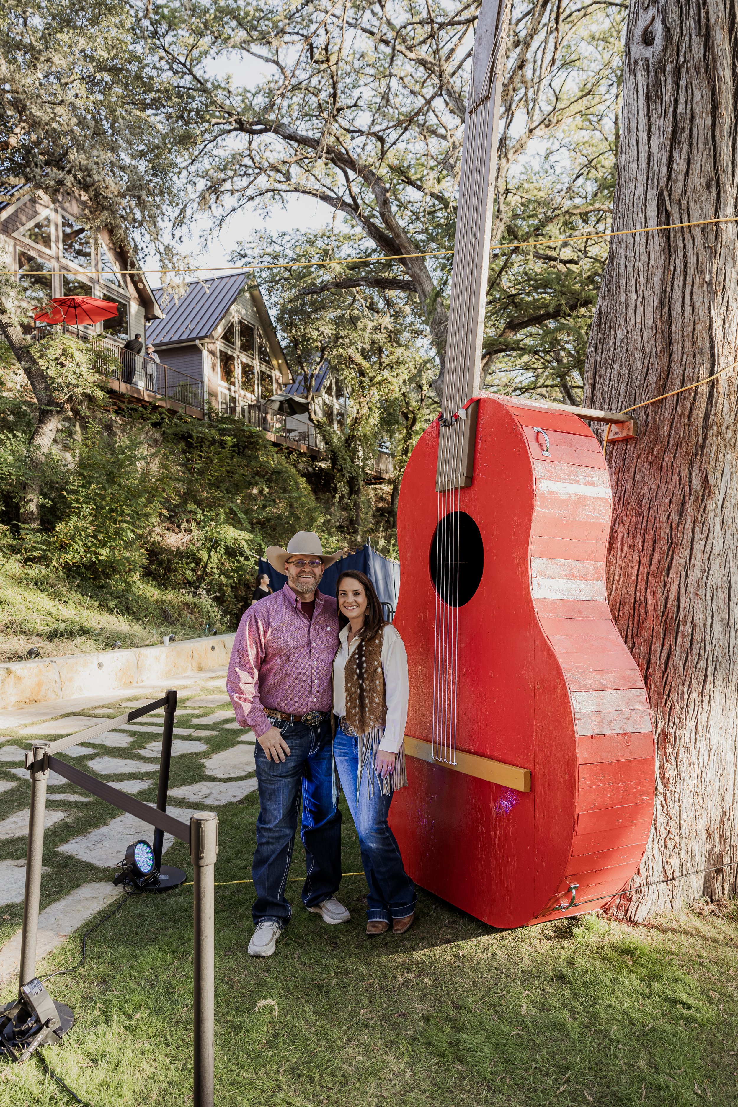 Two people, a man in a cowboy hat and a woman with long dark hair, standing and smiling next to a large red decorative guitar attached to a tree outdoors. There are houses on a hill in the background with large windows and a balcony with umbrellas.