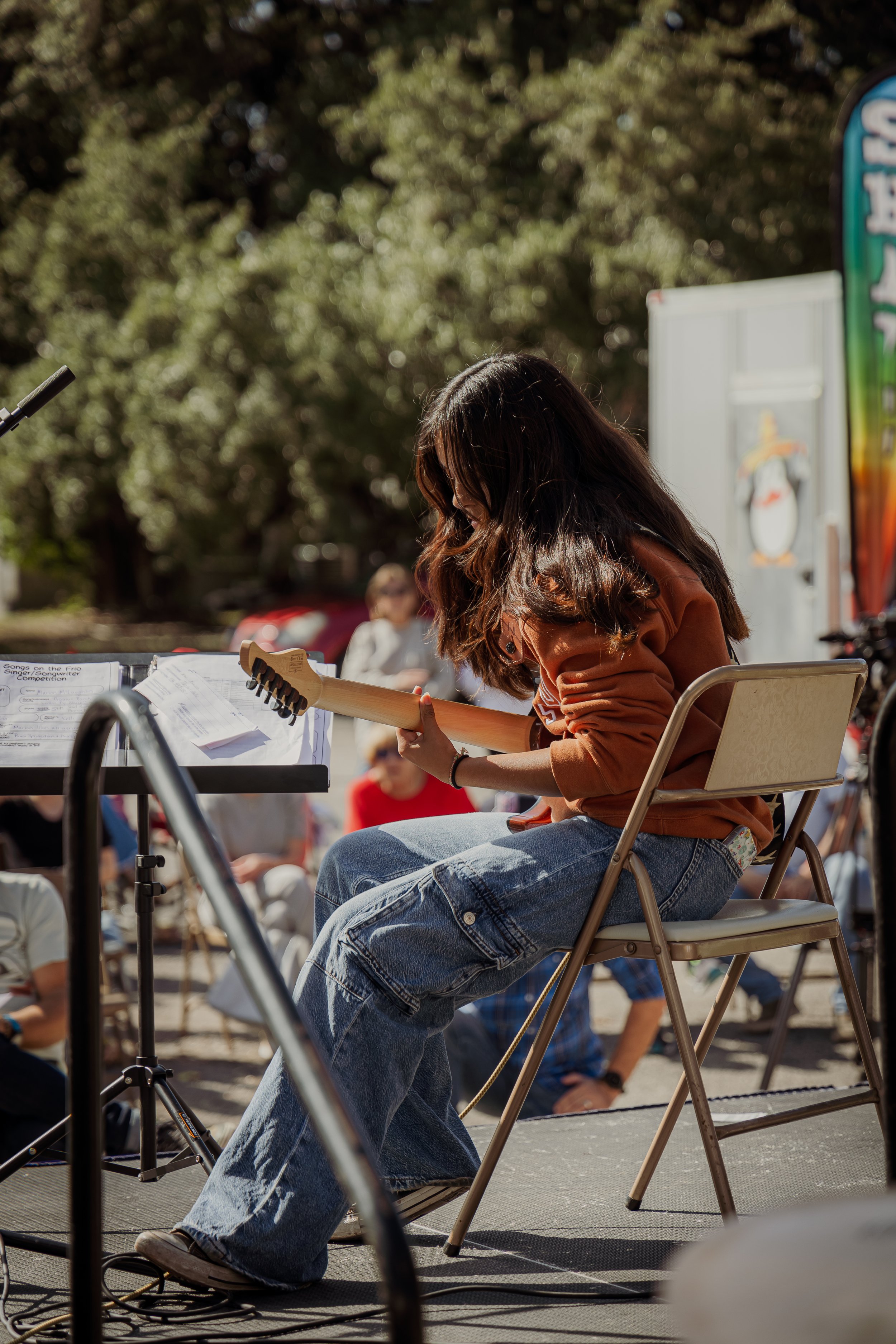 A woman with long dark hair, wearing a brown sweater and blue jeans, sitting on a folding chair and playing an electric guitar during an outdoor event.